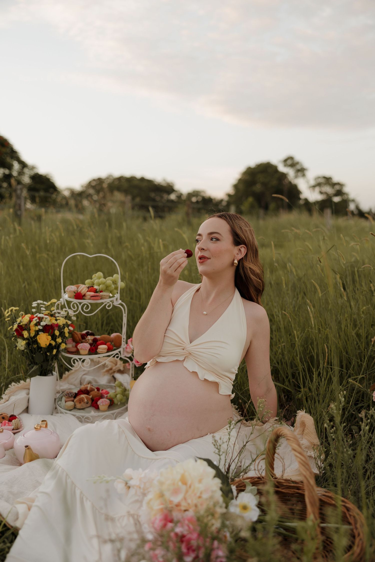 Pregnant woman sitting in a field enjoying a picnic with fruits, flowers, and treats on a blanket at sunset. bundaberg Photographer