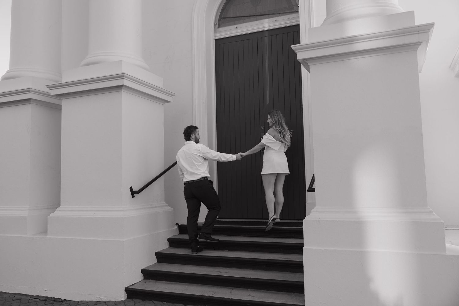 Black and white photo of a couple holding hands walking up steps to a grand white building entrance. Bundaberg Photographer