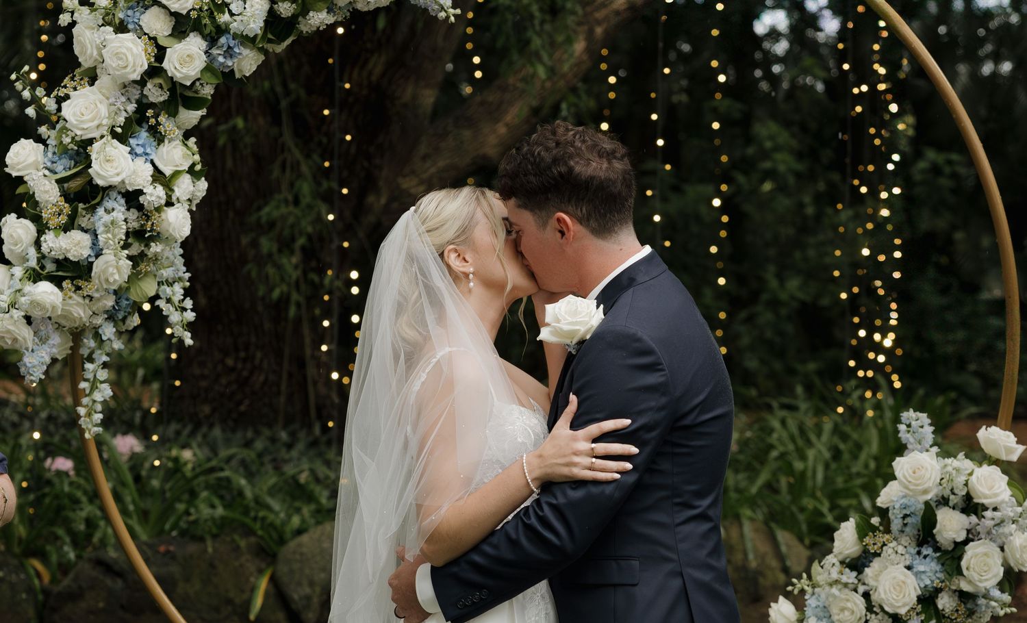 Bride and groom share first kiss under floral arch with fairy lights at outdoor wedding ceremony. Bundaberg photographer