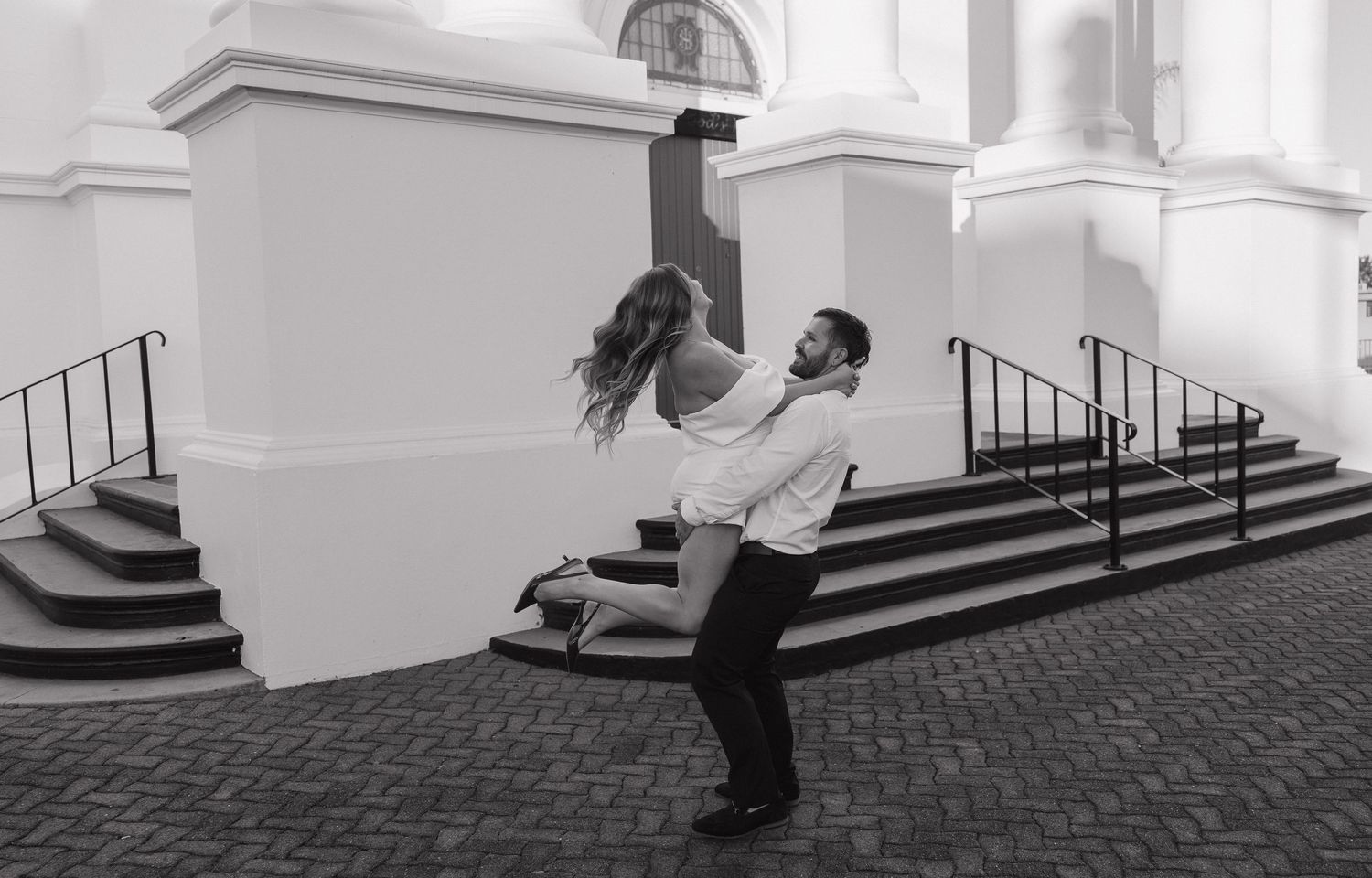 Black and white photo of a man lifting a woman outdoors near classic white building steps in a romantic dance pose. Bundaberg photographer