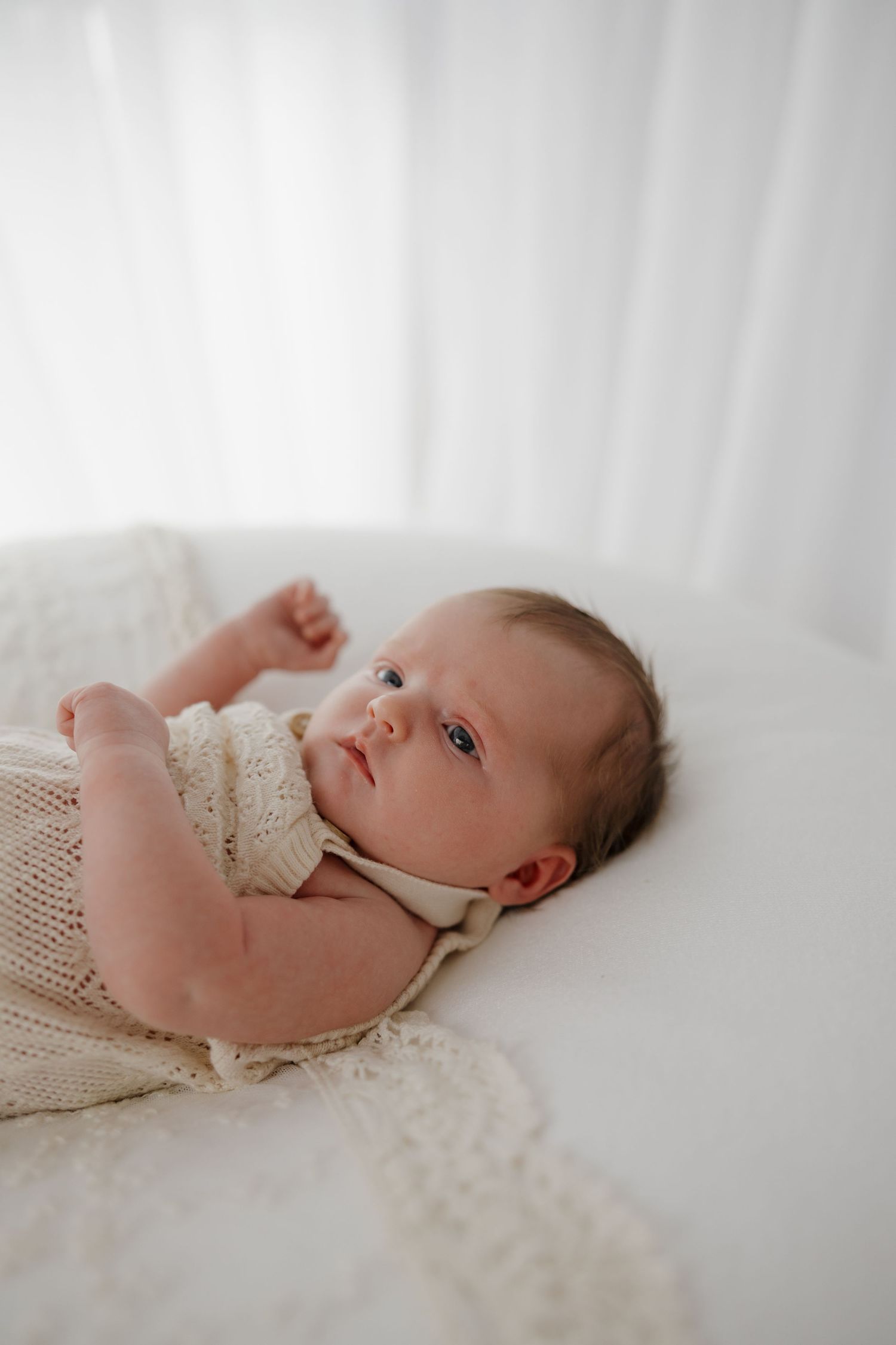 A peaceful newborn baby in a cream knit romper lying on a white blanket, gazing upward with bright blue eyes. Bundaberg photographer