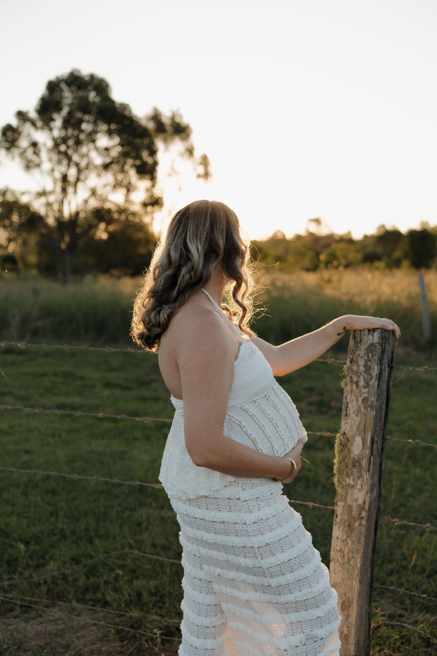Pregnant woman in white lace dress holding baby bump at golden hour by rustic fence in green field. Bundaberg photographer