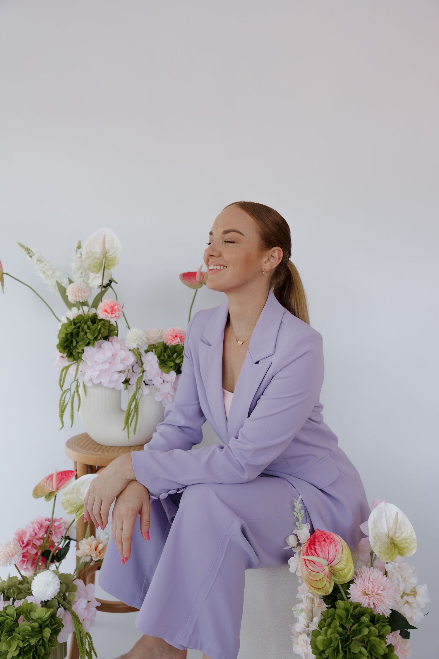 Woman in lavender suit sitting and smiling among pink and white flowers against a white background. Bundaberg photographer
