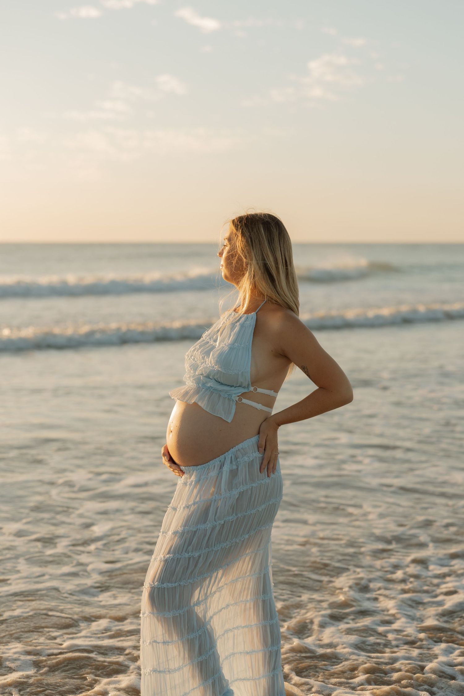 Pregnant woman in white outfit cradling her baby bump on a sandy beach at golden hour sunset. Bundaberg Photographer