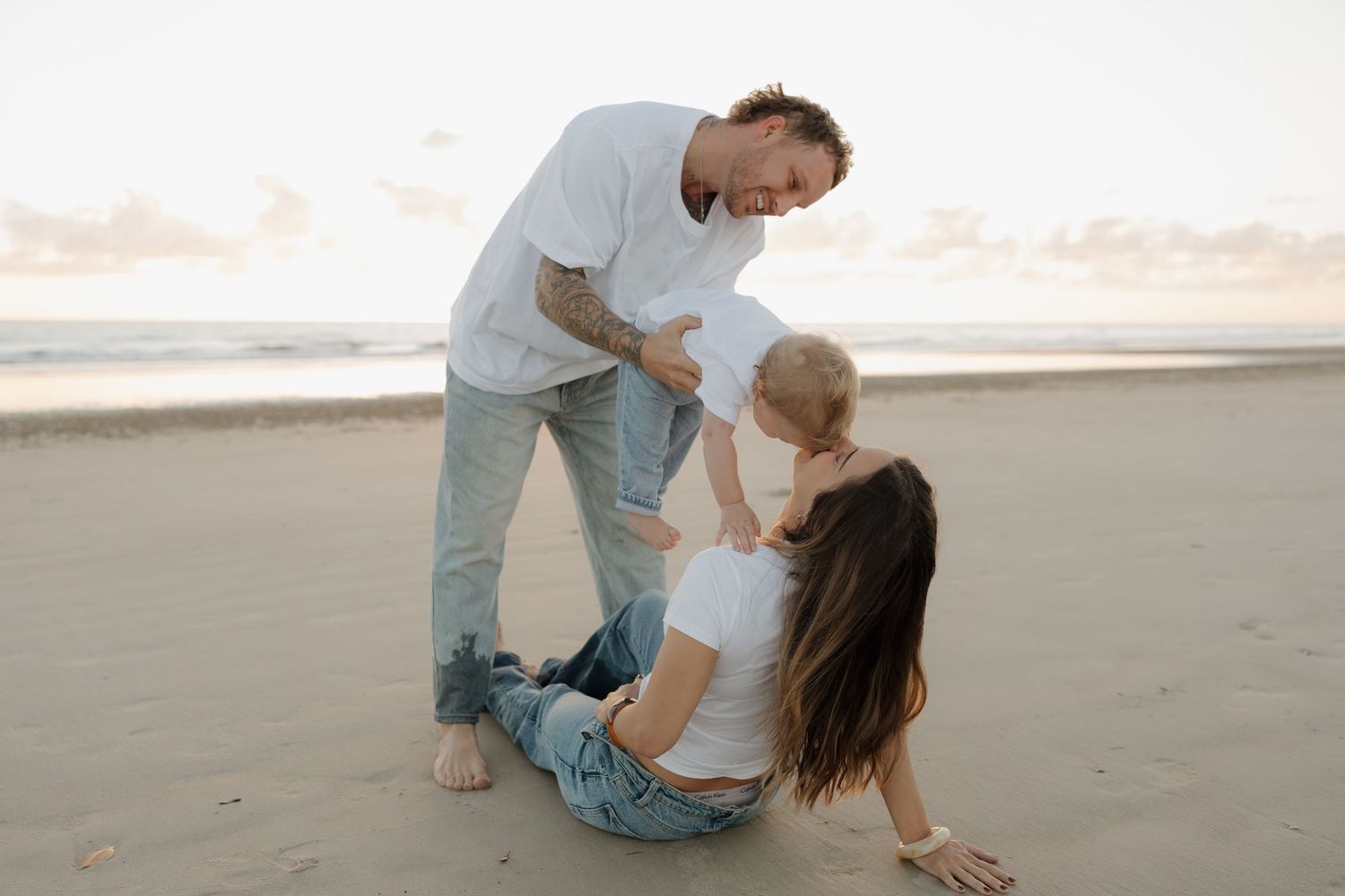 Family of three enjoying a beach day, with parents playing with their baby on sandy shore at sunset. Bundaberg Photographer