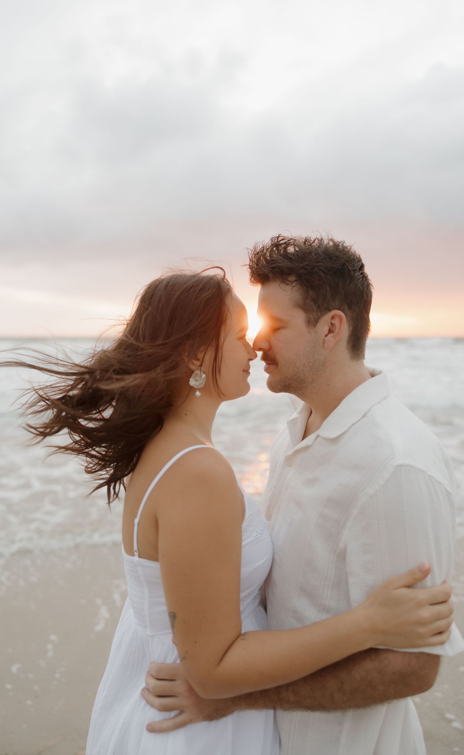 Romantic couple in white outfits embracing on a beach at sunset, with ocean waves and a glowing sky behind them. Bundaberg photographer