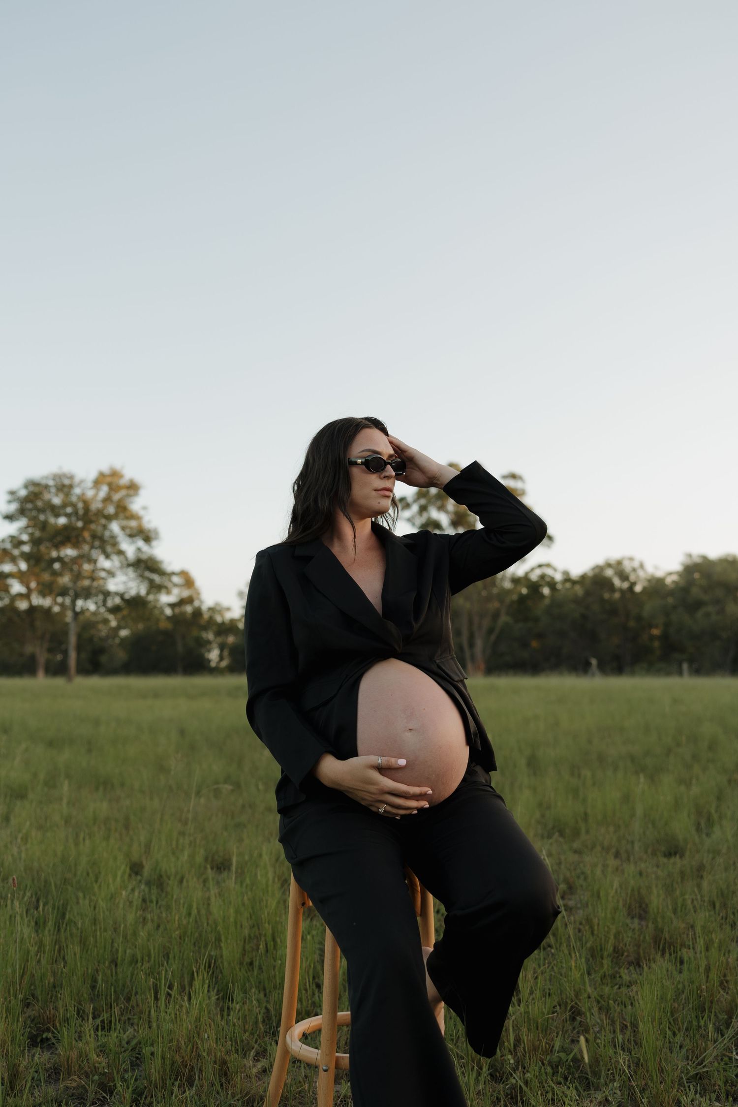 Pregnant woman in black suit posing outdoors on stool in green field, cradling baby bump, wearing sunglasses at dusk. Bundaberg photographer