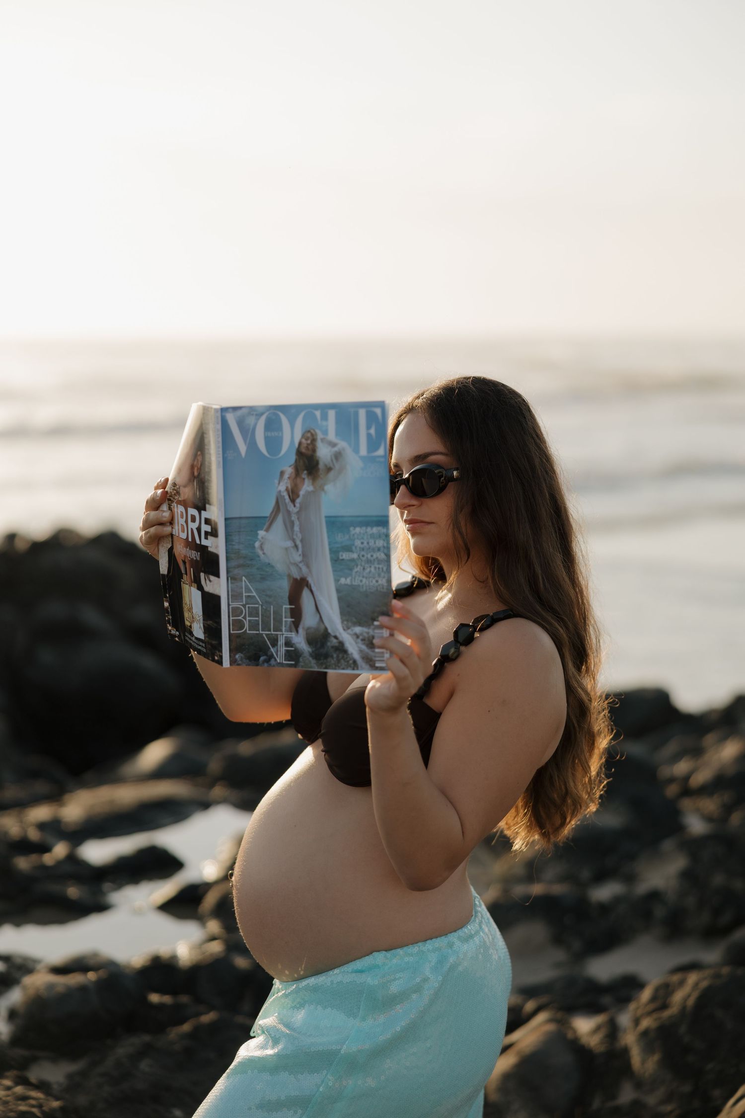 Pregnant woman in black bikini top reads Vogue magazine on rocky beach at sunset wearing sunglasses. Bundaberg Photographer