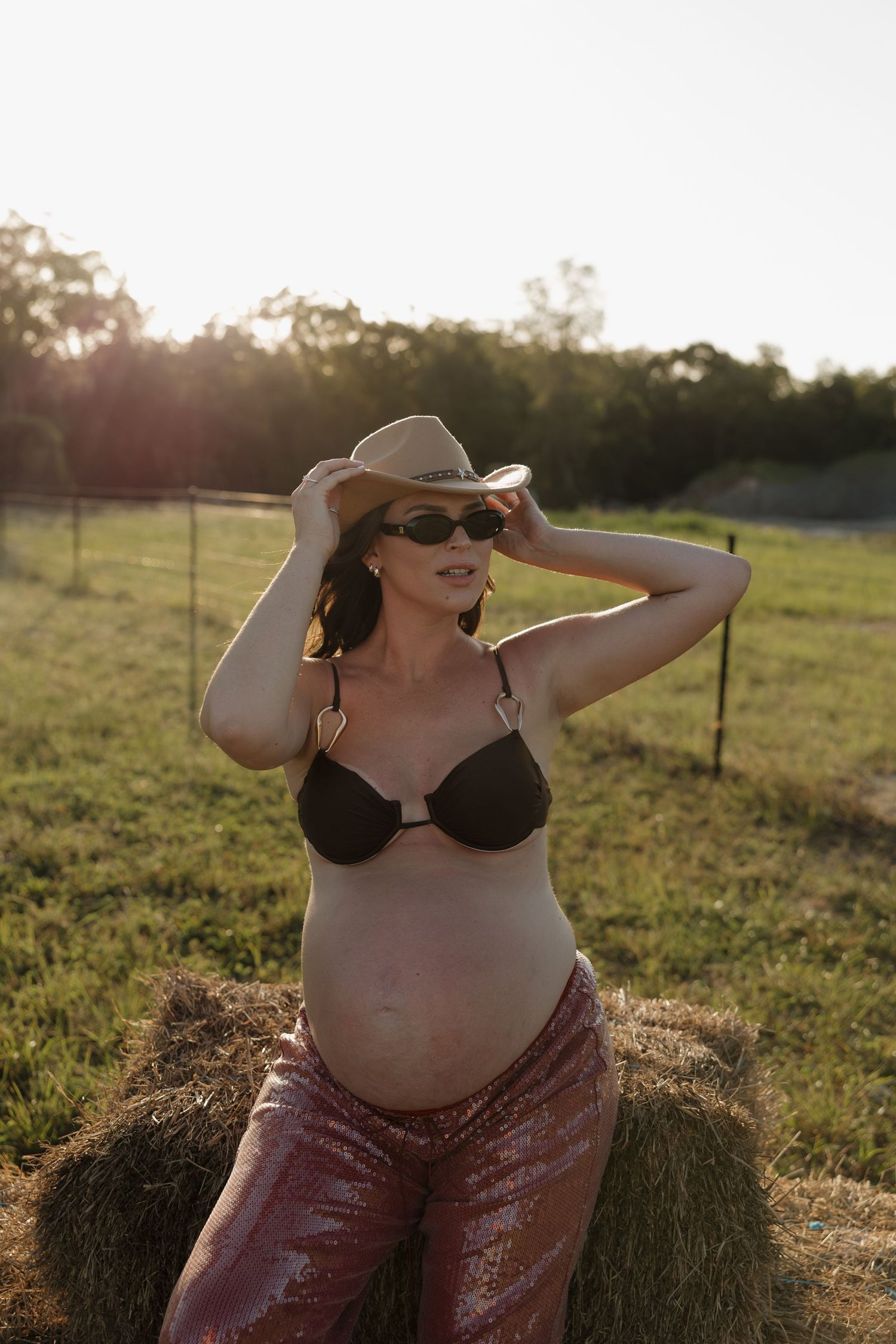Pregnant woman in black bikini top, sequin pants, cowboy hat, and sunglasses posing by hay bale in sunny field. Bundaberg photographer