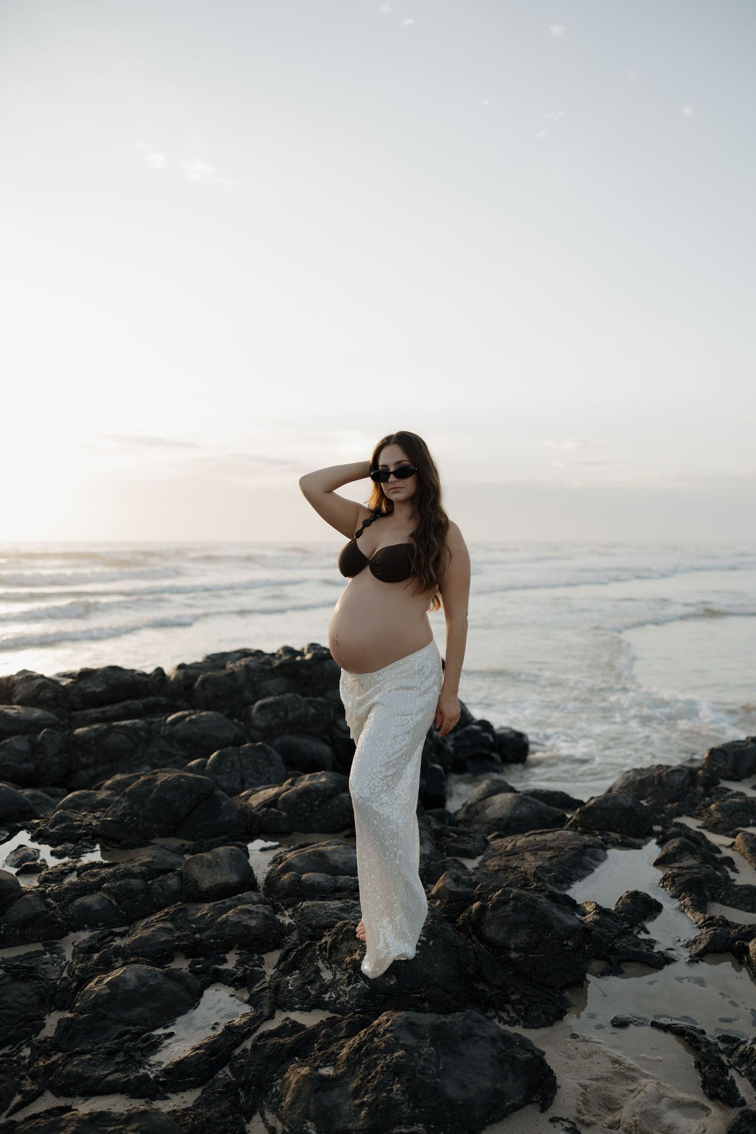 Pregnant woman in black bikini top and white skirt poses on rocky beach at sunset wearing sunglasses. Lovely photography bundaberg photo