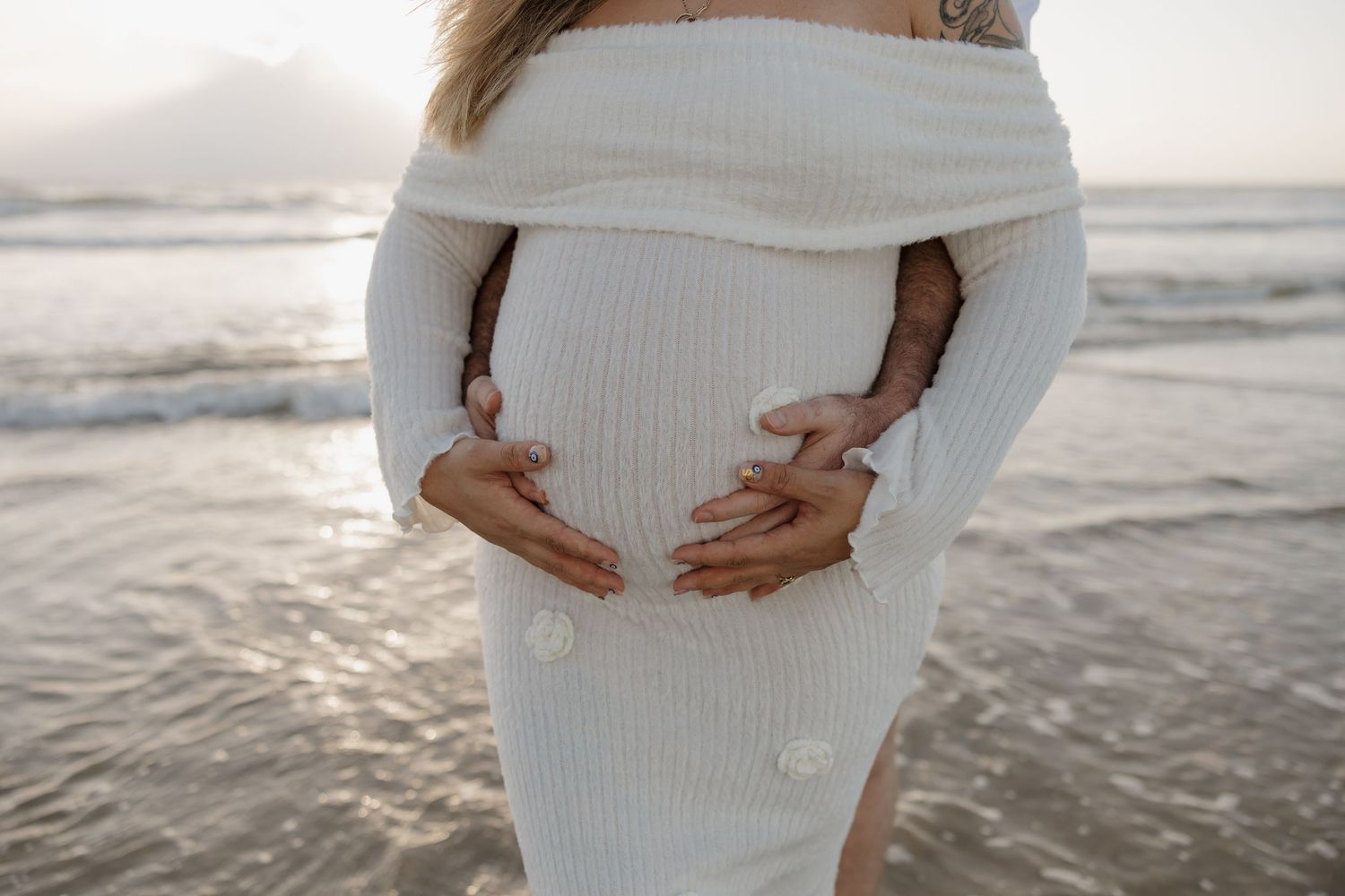 Pregnant woman in white ribbed off-shoulder dress cradling baby bump on a serene beach at sunset. Bundaberg photographer