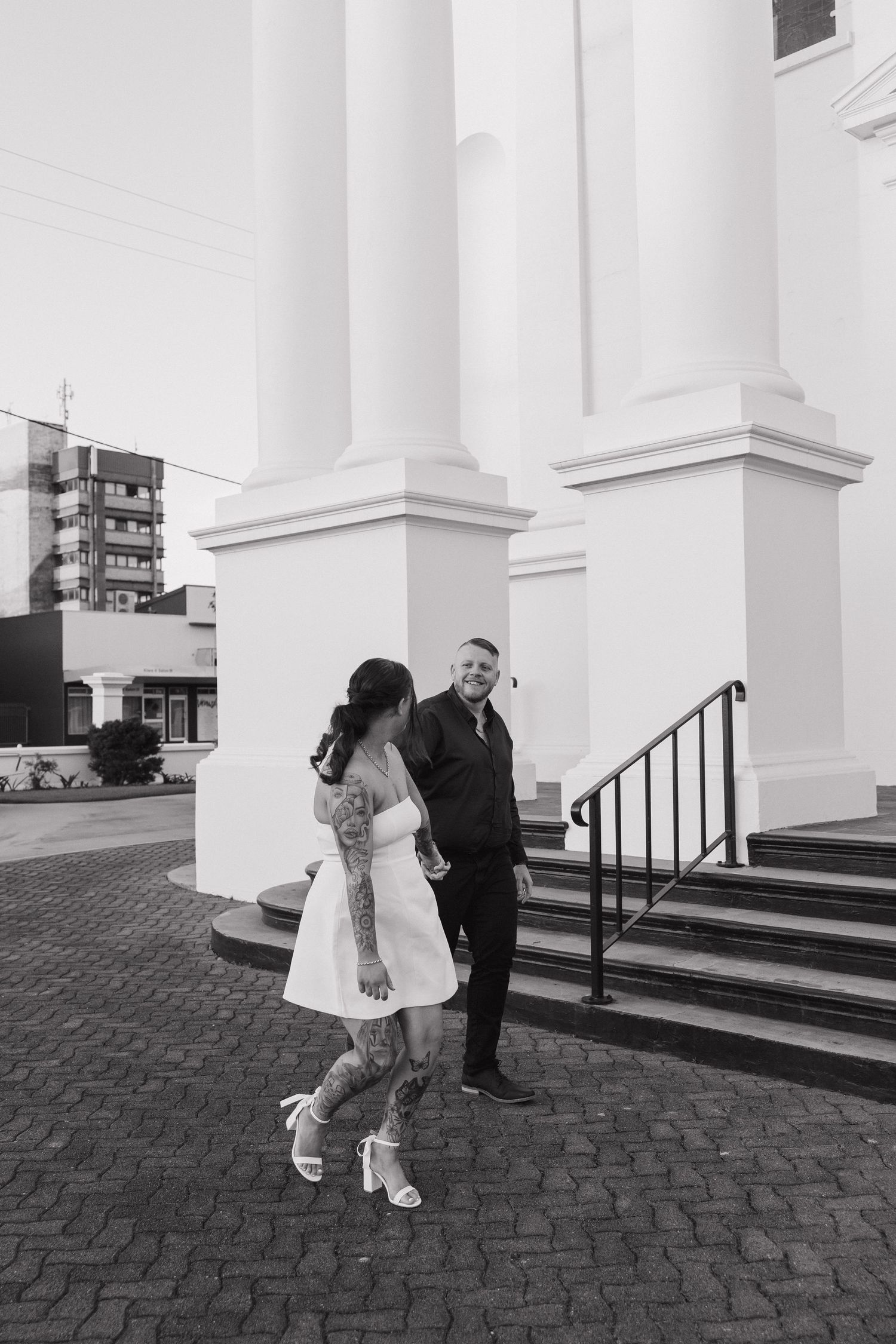Black and white photo of a tattooed bride in white dress and groom in black suit laughing outside a white church. Bundaberg photographer
