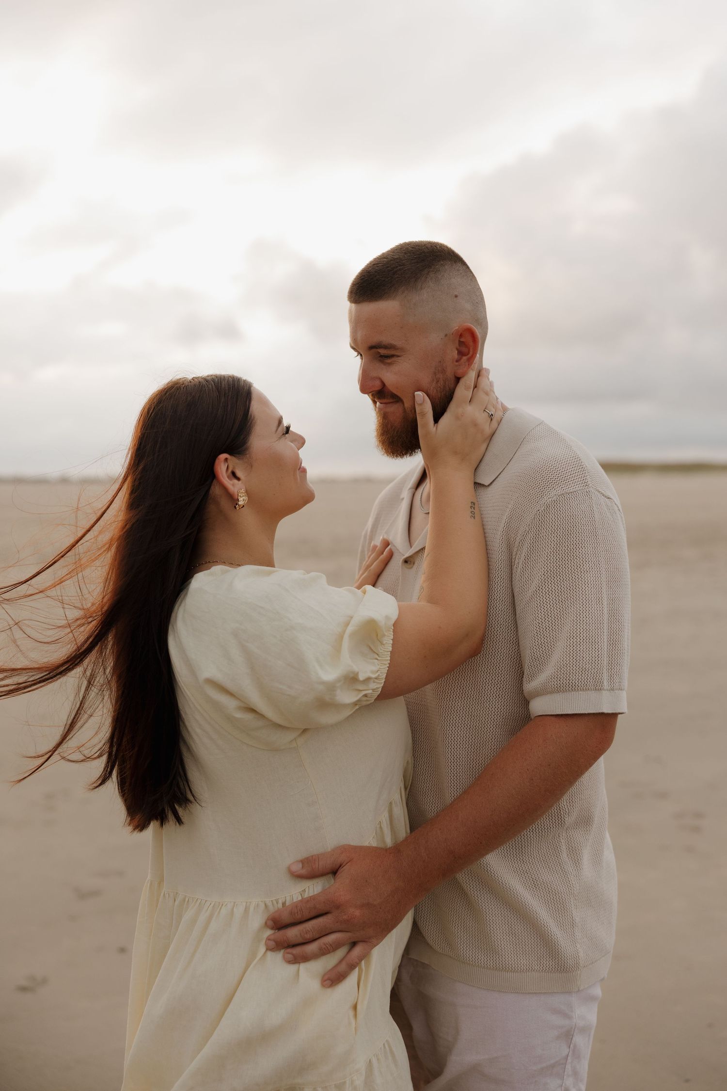 A couple embraces on a sandy beach, the woman touching the bearded man's face as they smile at each other. Bundaberg photographer