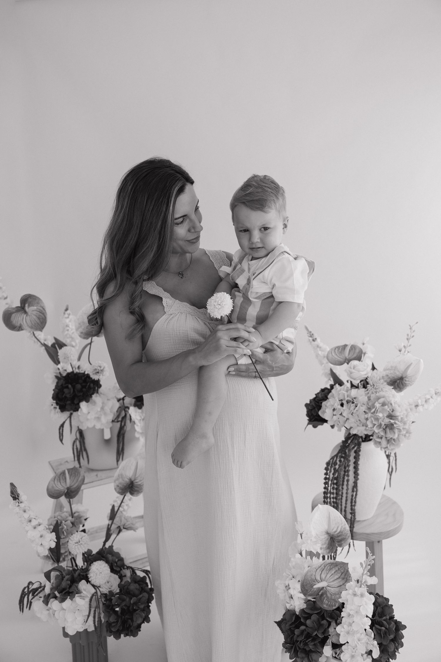 Black and white photo of a mother in white dress holding her toddler surrounded by floral arrangements. Bundaberg photographer