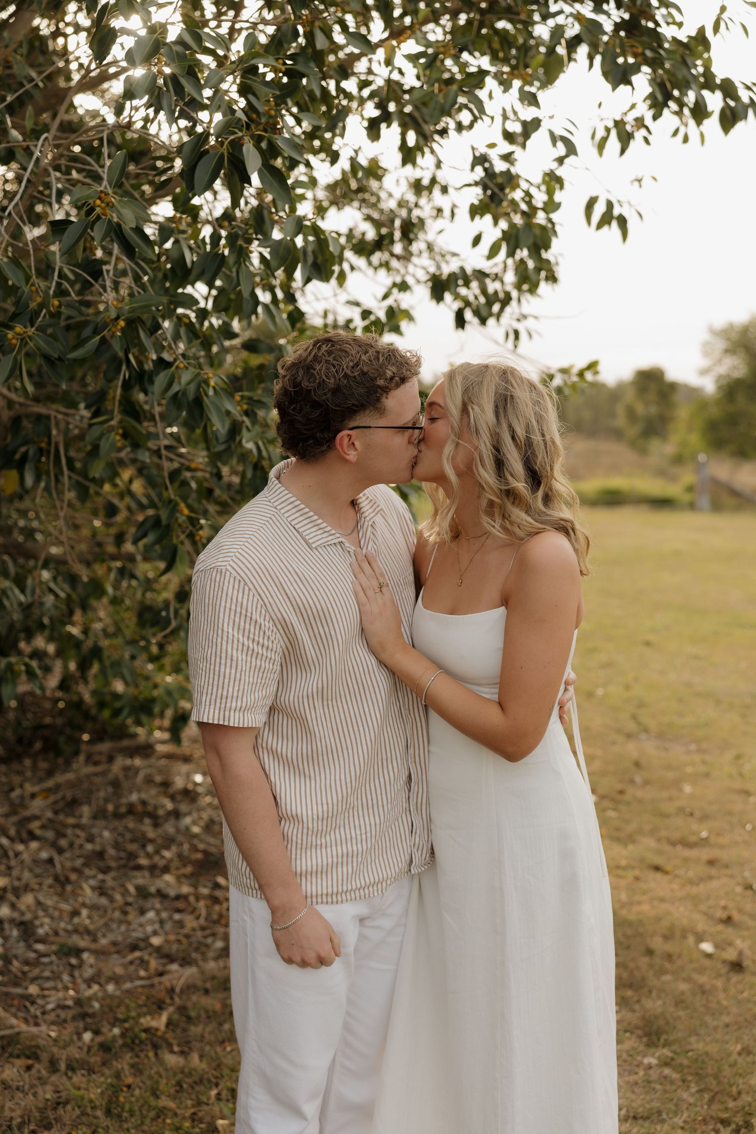 Couple sharing a romantic kiss outdoors near a leafy tree, wearing white and striped outfits in a sunlit garden. Bundaberg photographer