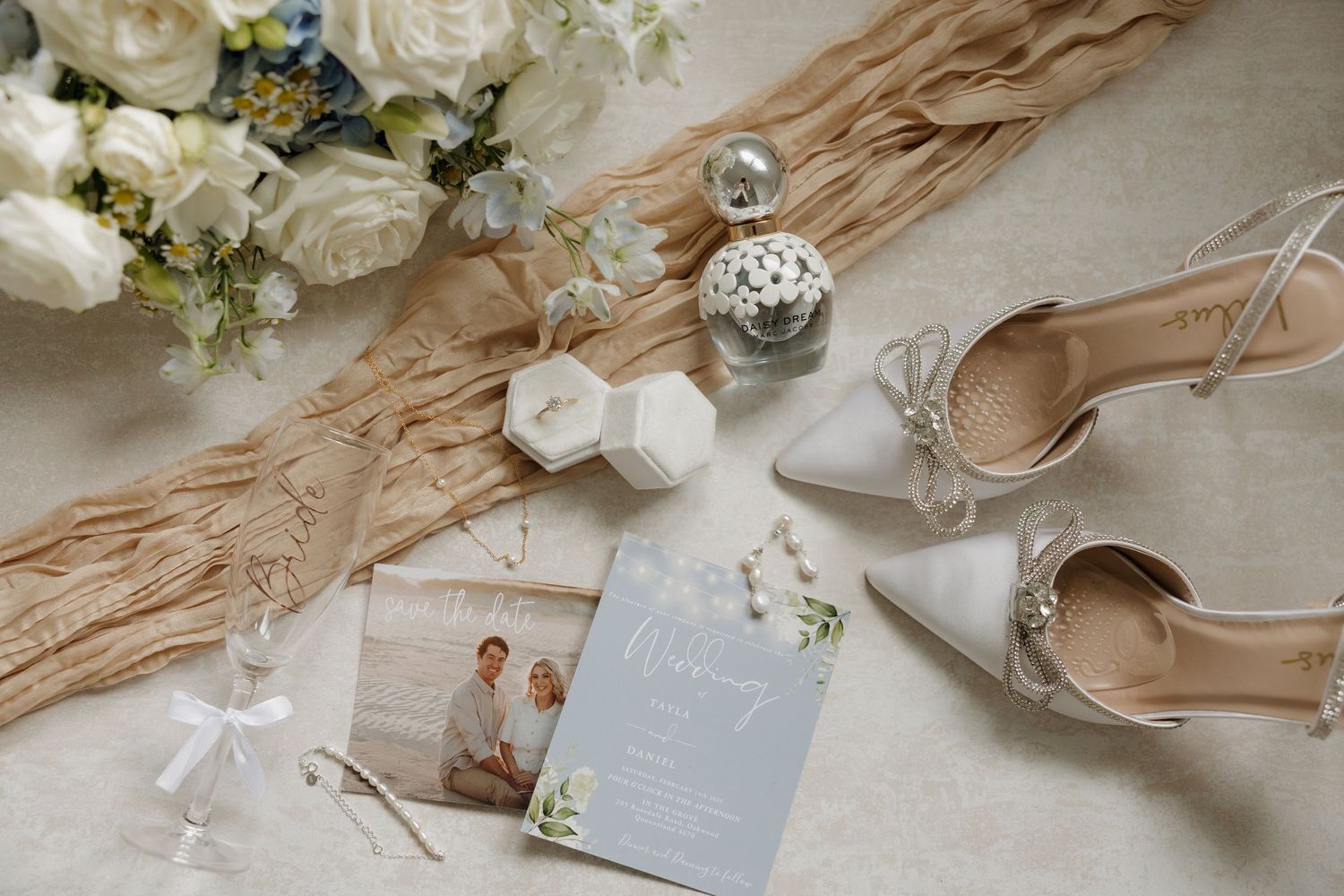 Elegant wedding flat lay with white heels, floral bouquet, invitation, perfume, and ring box on neutral background. Bundaberg photographer