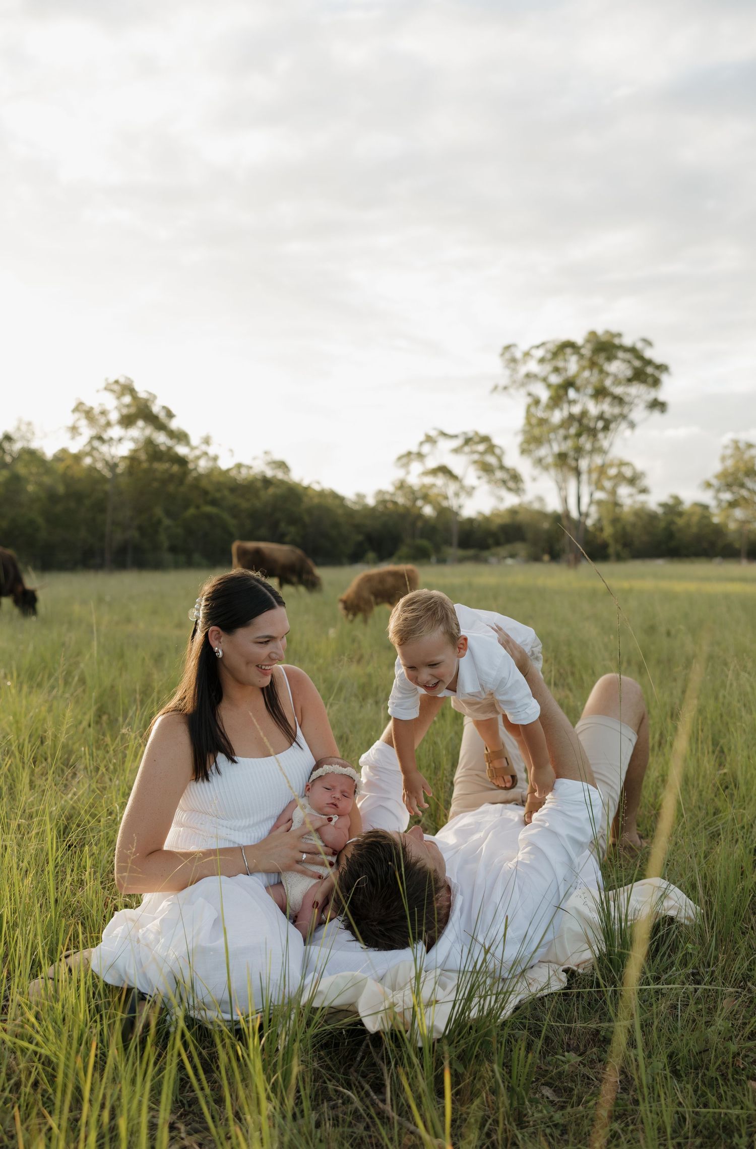 Family in white clothing sitting in green field, mother holding newborn baby while father lifts toddler outdoors. Bundaberg photographer