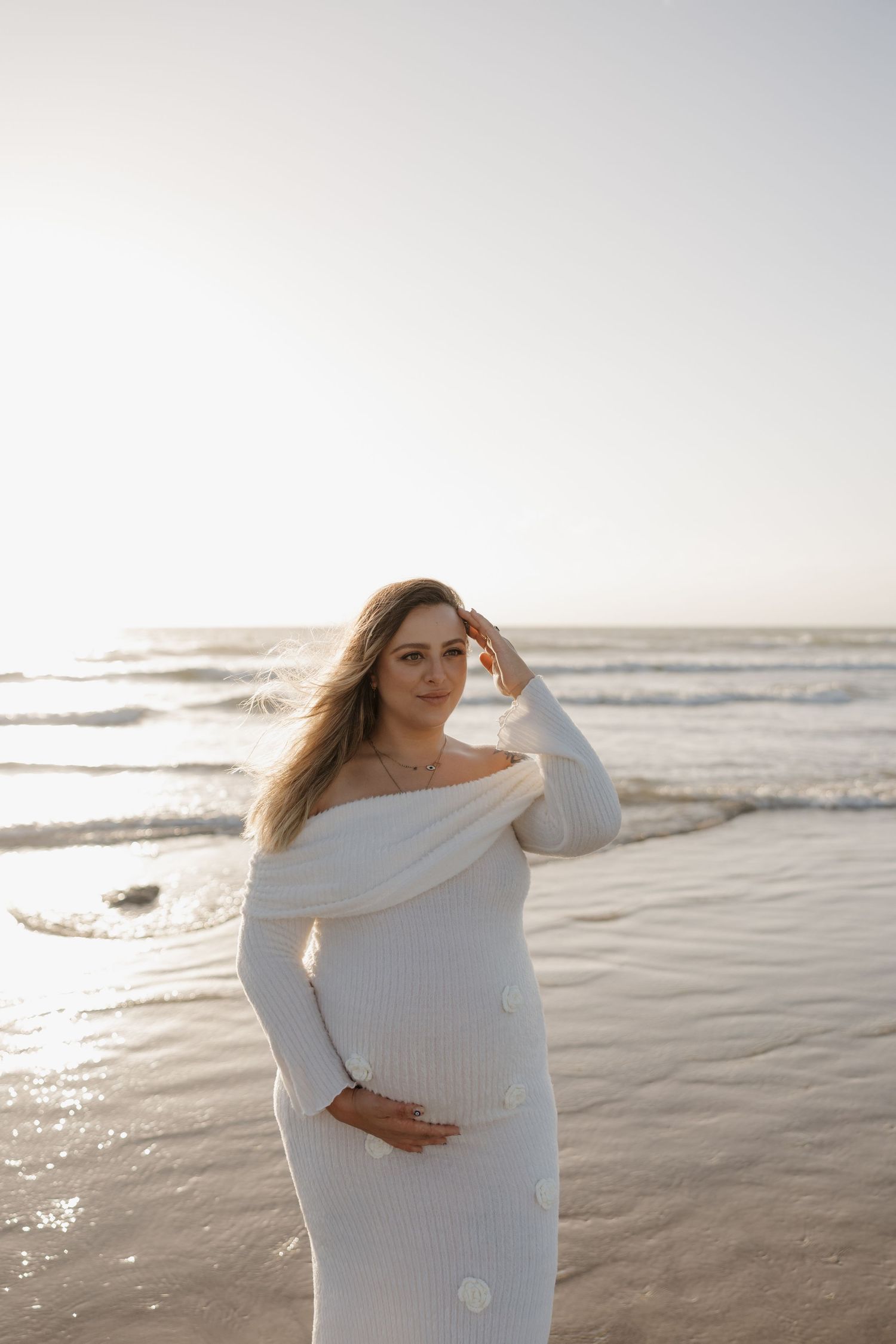 Pregnant woman standing on beach at sunset, cradling baby bump with ocean waves behind her. Bundaberg Photographer