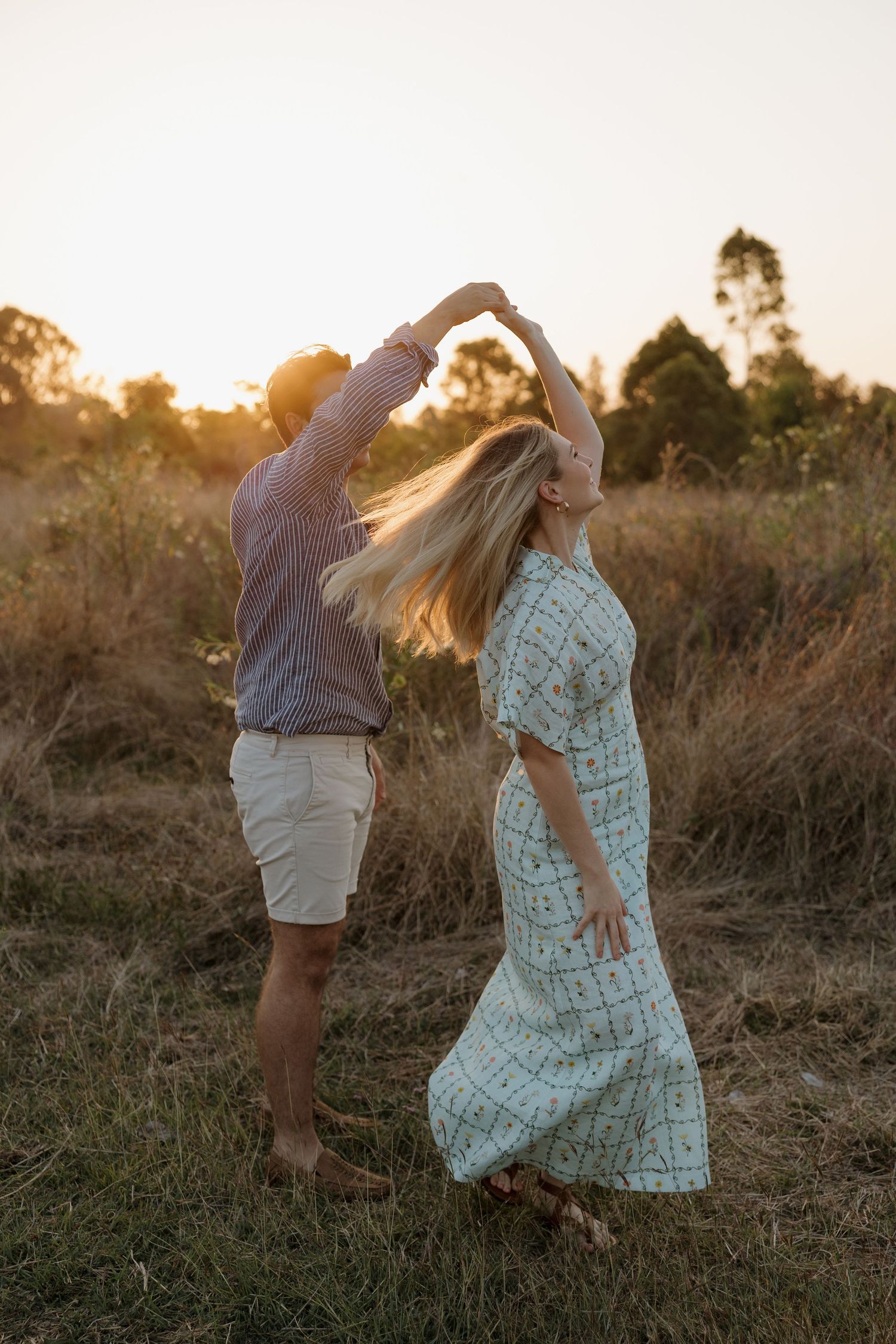 Couple dancing outdoors at golden sunset in a field, woman in floral maxi dress twirling with flowing hair. Bundaberg Photographer