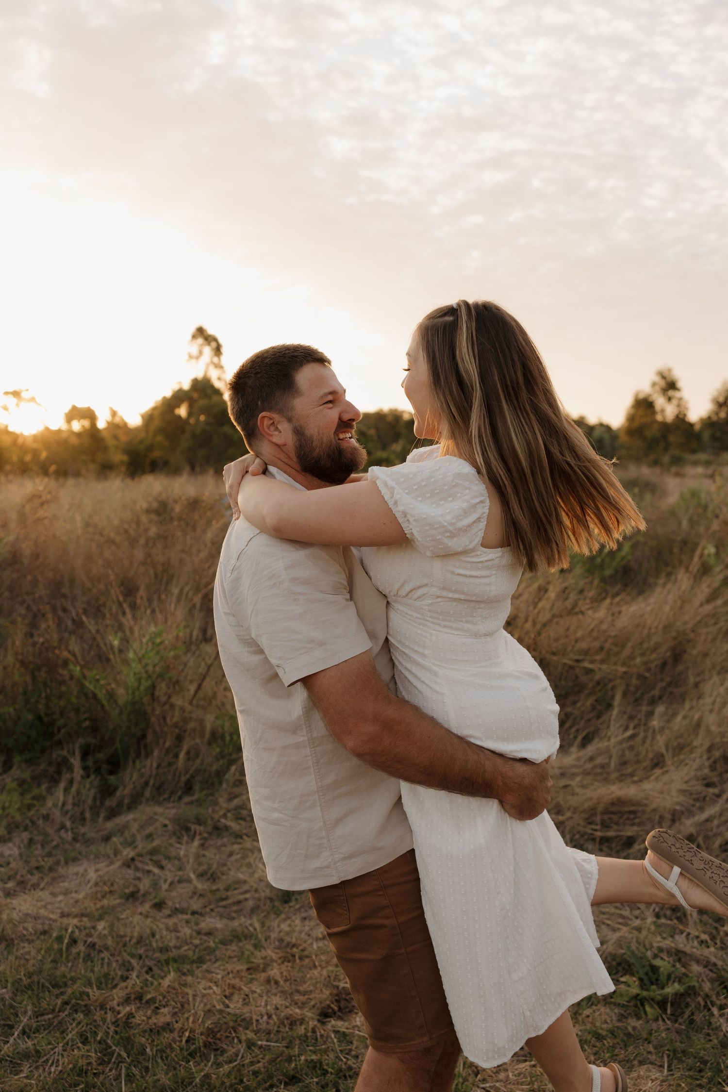 A couple together in a golden field at sunset, the woman in a white dress lifting her leg joyfully. Bundaberg Photographer