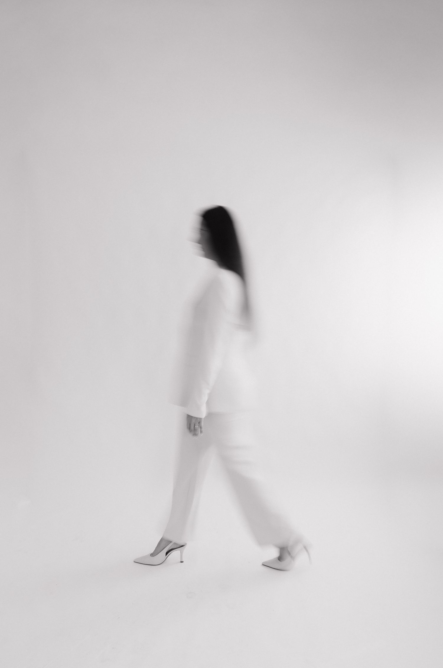 Black and white motion blur photo of a woman in white outfit and heels walking against a light background. Bundaberg photographer