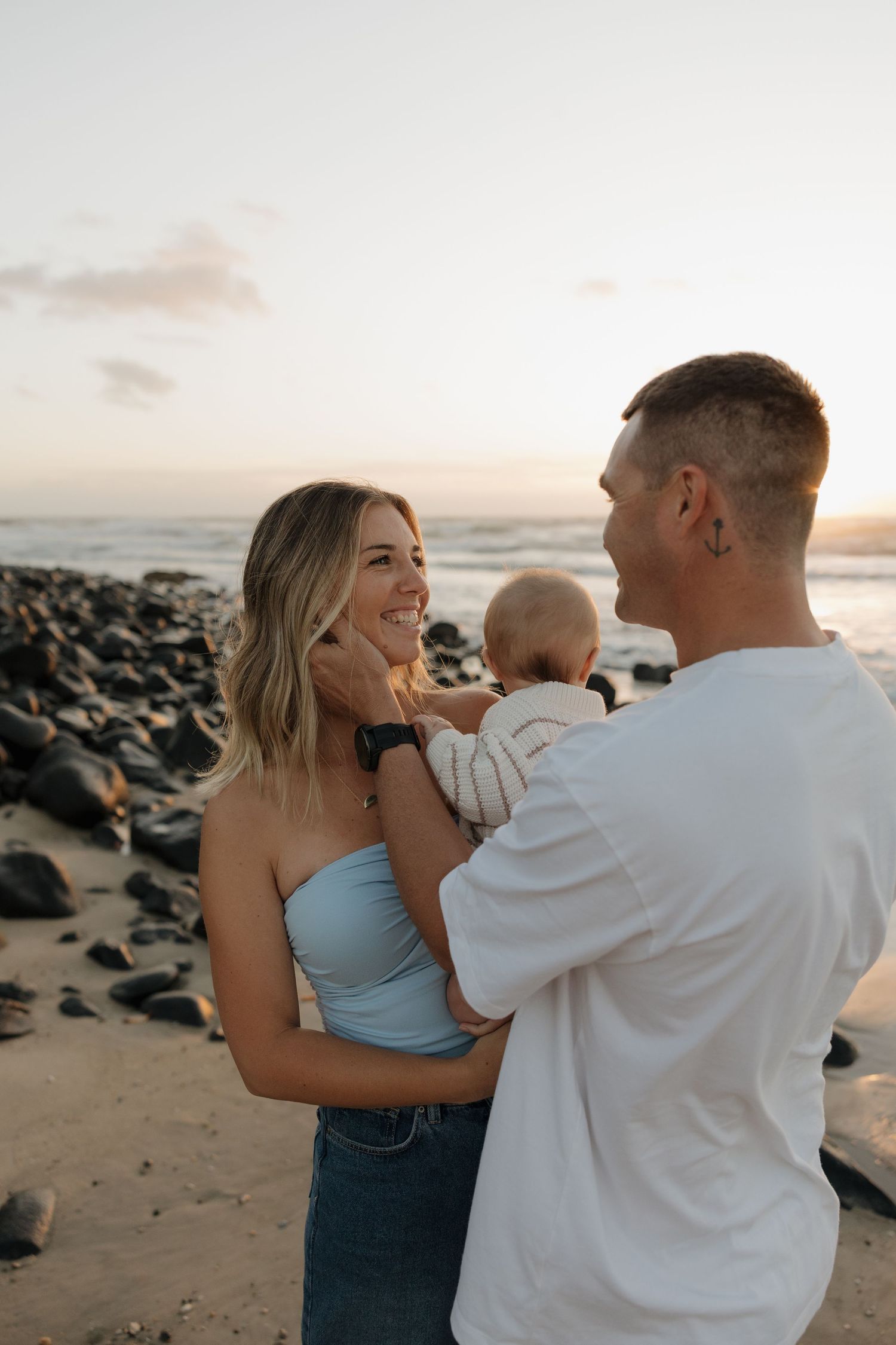 A young couple smiles at each other while holding their baby on a rocky beach at sunset. Bundaberg photographer