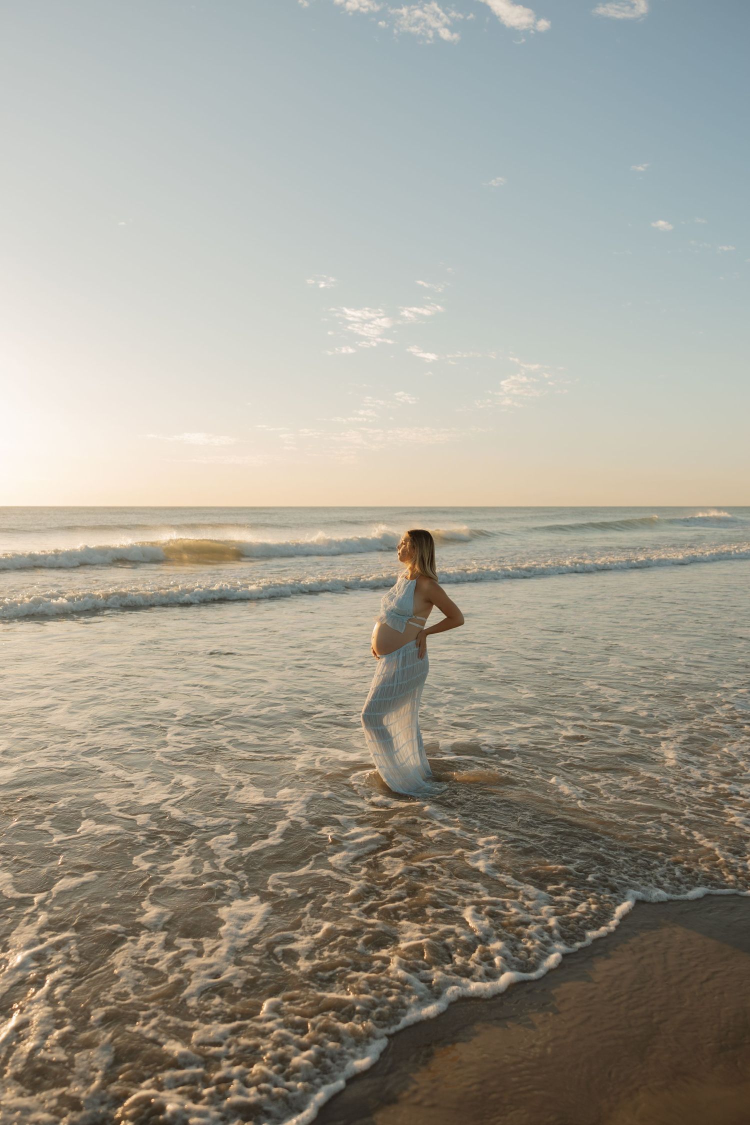 Pregnant woman in white dress standing in ocean waves at sunset on a sandy beach. Bundaberg Photographer
