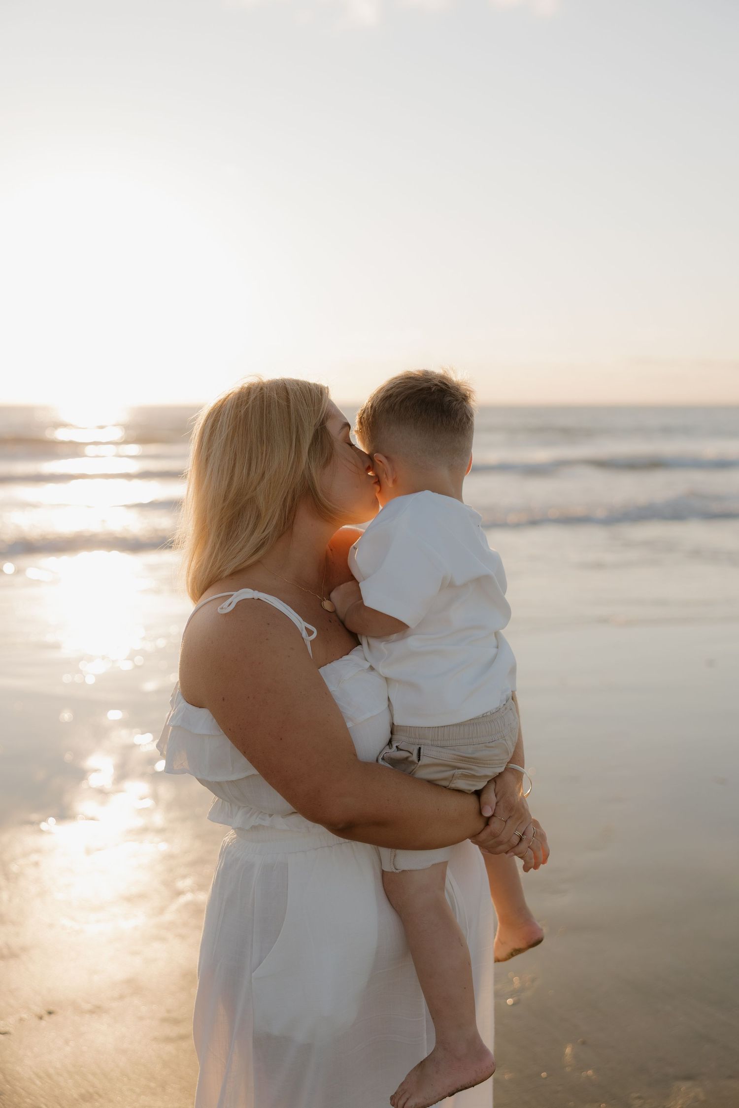 A mother in a white dress holds her toddler on a sunlit beach, sharing a tender kiss at golden hour. Bundaberg Photographer