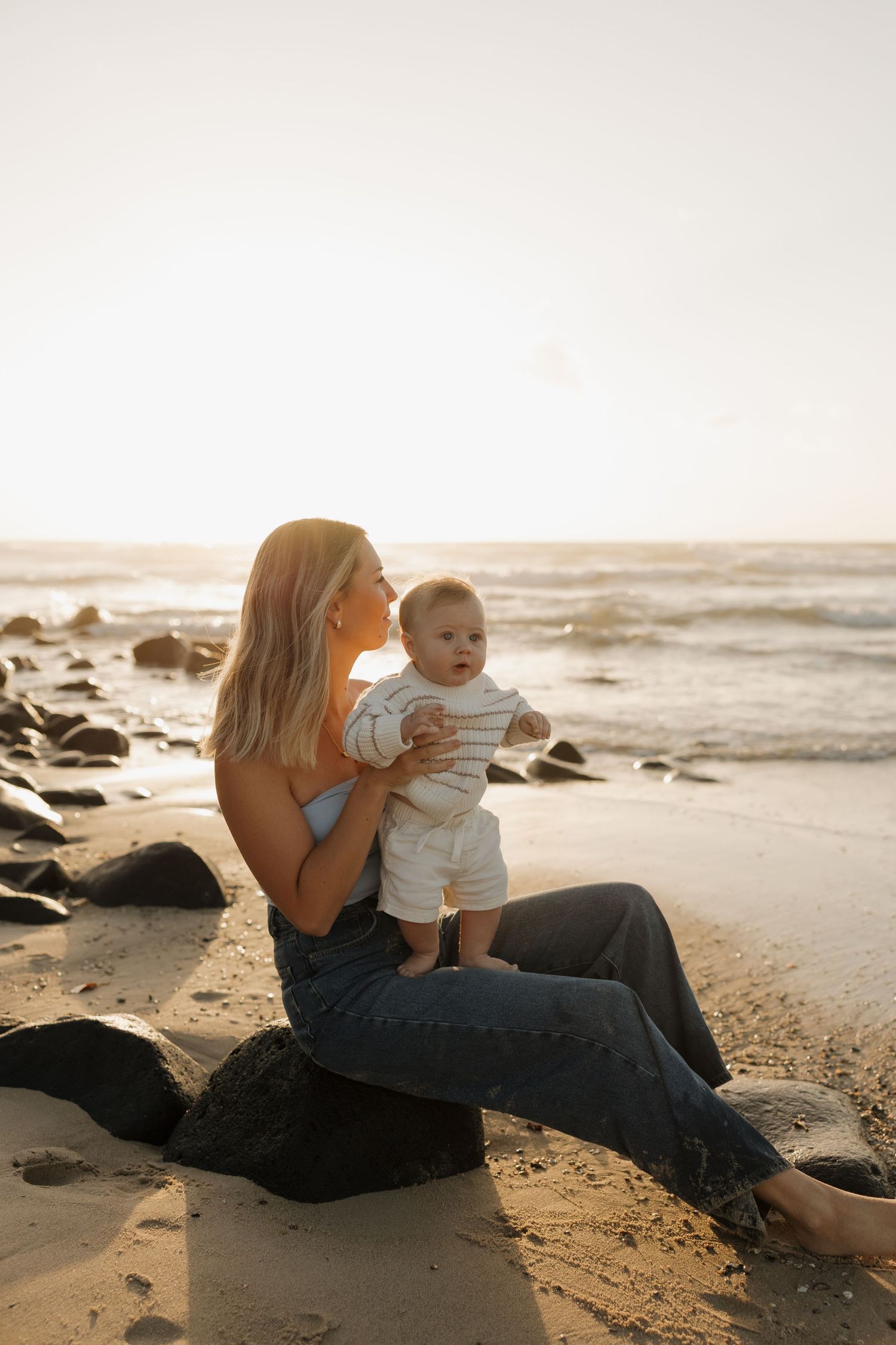 A mother sits on a rock at the beach during golden hour, holding her baby while ocean waves roll in behind them. Bundaberg photographer