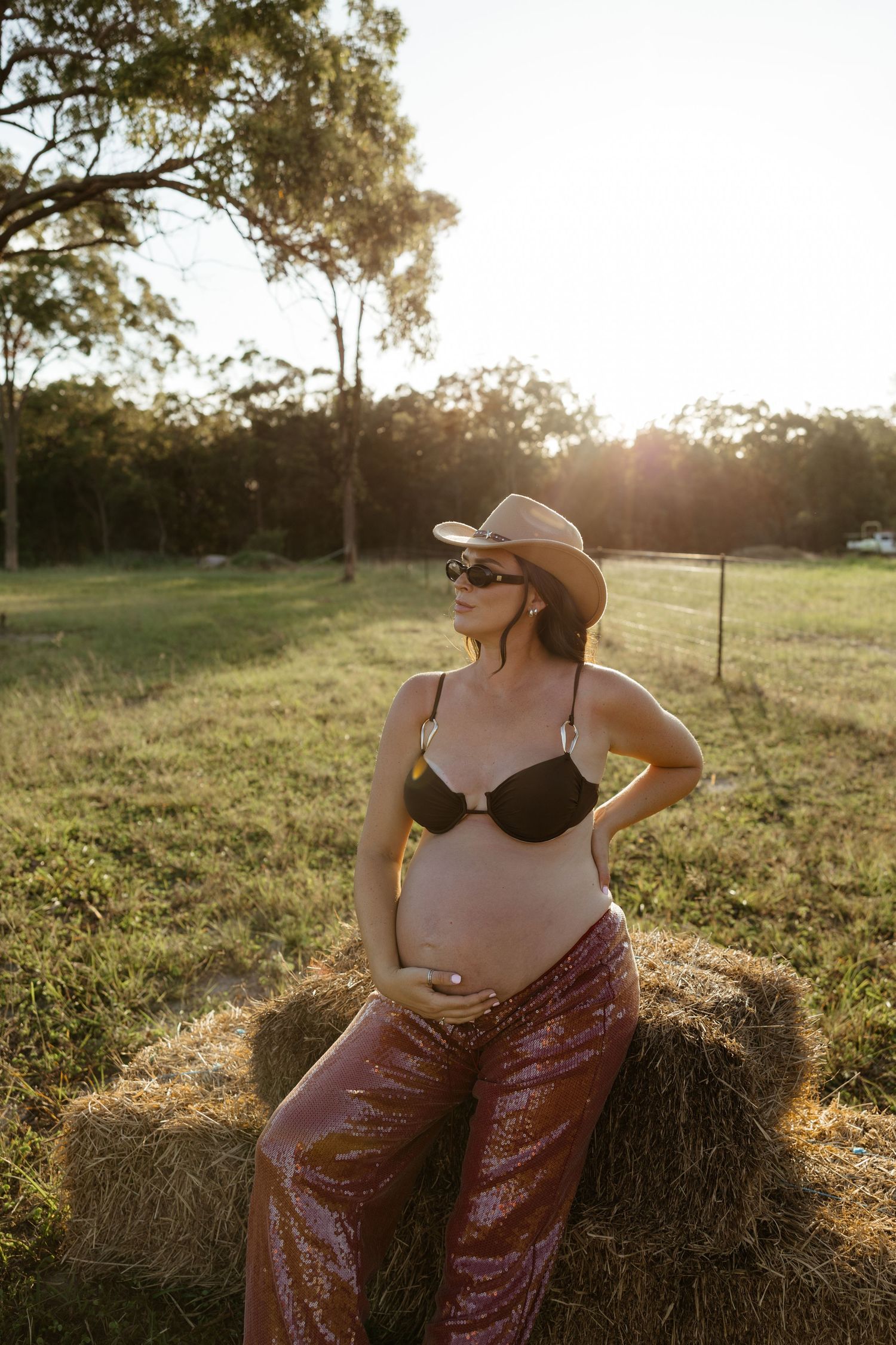 Pregnant woman in cowboy hat, sunglasses, and sequin pants leaning on hay bale at golden hour. Bundaberg Photographer