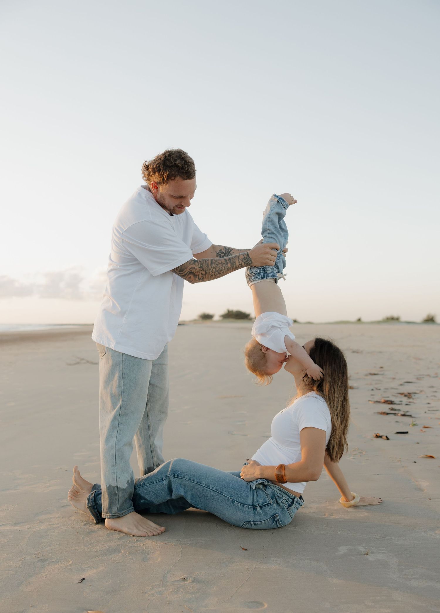 A tattooed father holds a toddler upside down while a mother sits on a sandy beach, all wearing white tops and jeans. Bundaberg Photographer