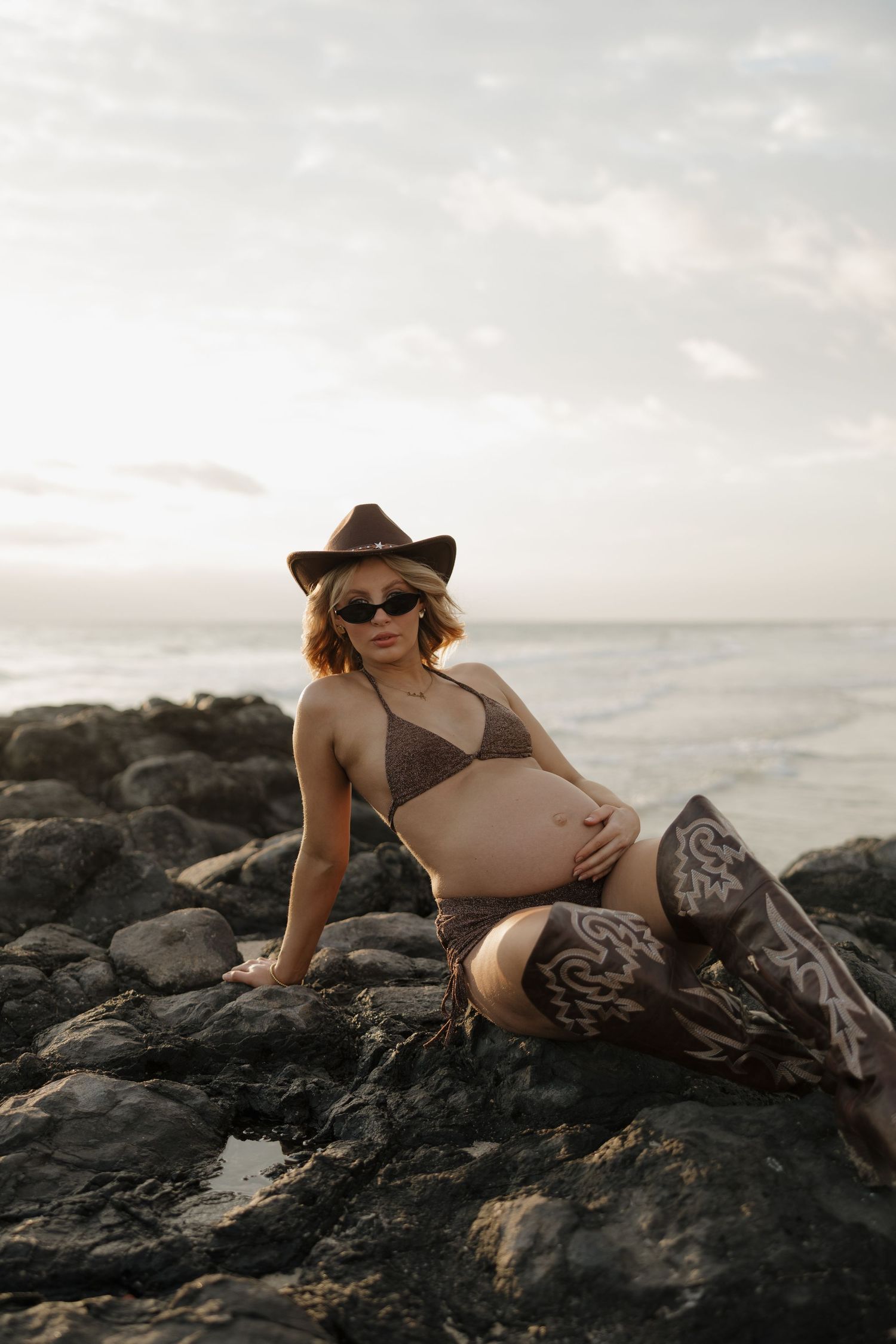 Pregnant woman in brown bikini, cowboy hat, sunglasses, and boots posing on rocky shoreline at sunset.Bundaberg photographer maternity vogue