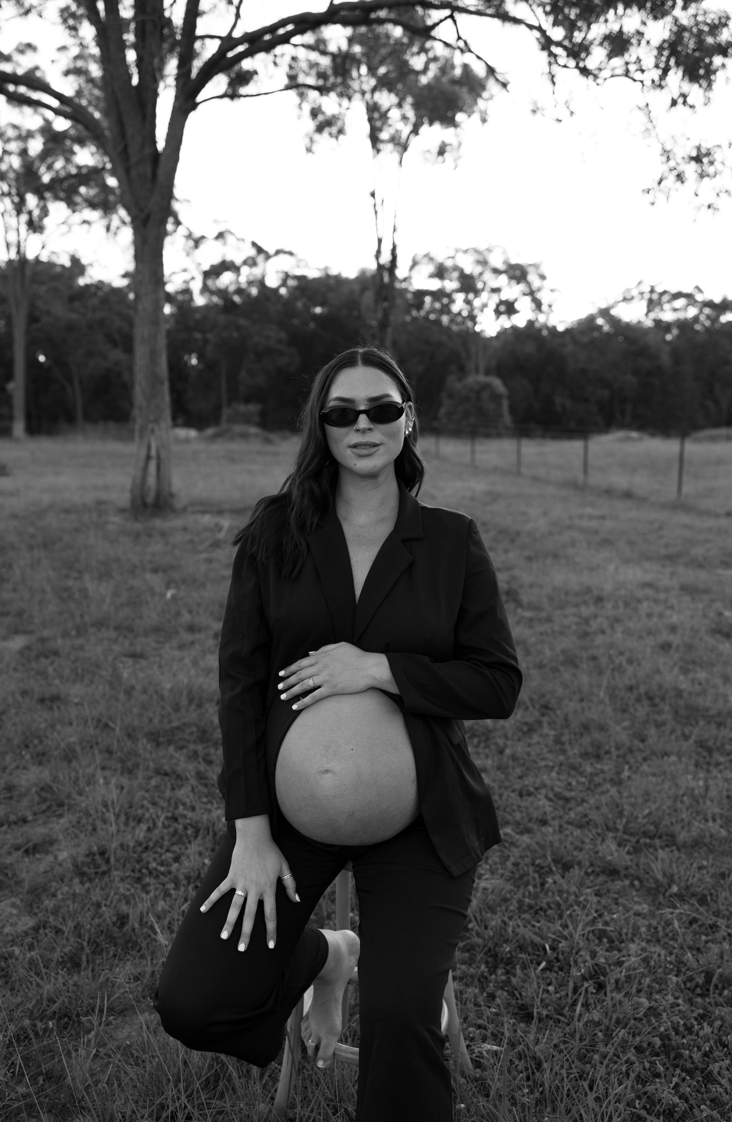 Pregnant woman in black outfit and sunglasses poses outdoors in a field with trees in this elegant maternity photo. Bundaberg photographer