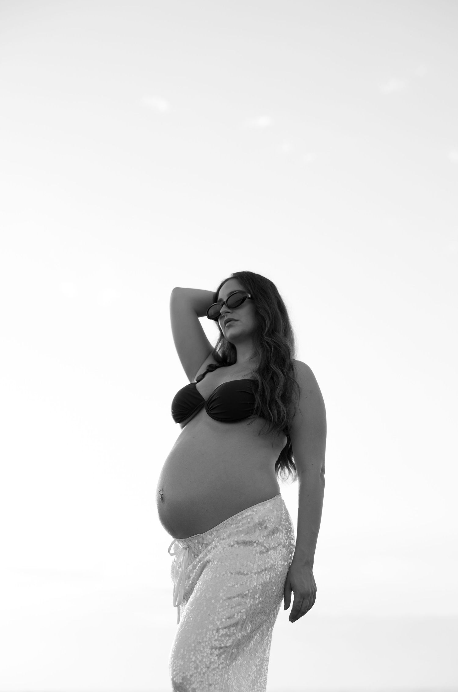 Black and white maternity photo of a pregnant woman in a bikini top and skirt, posing confidently outdoors. Bundaberg Photographer