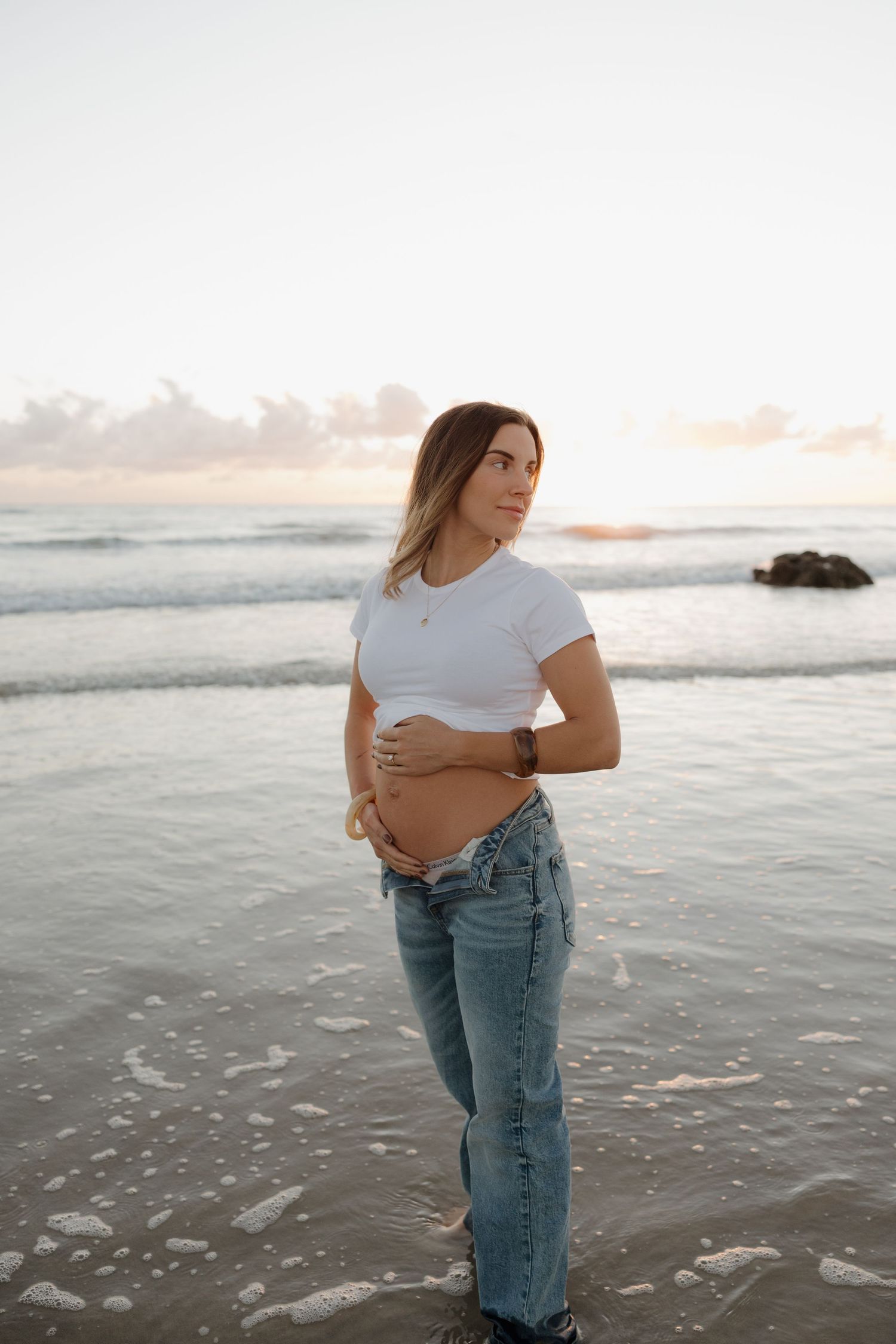 Pregnant woman in white crop top and jeans cradling baby bump on beach at sunset. Bundaberg Photographer