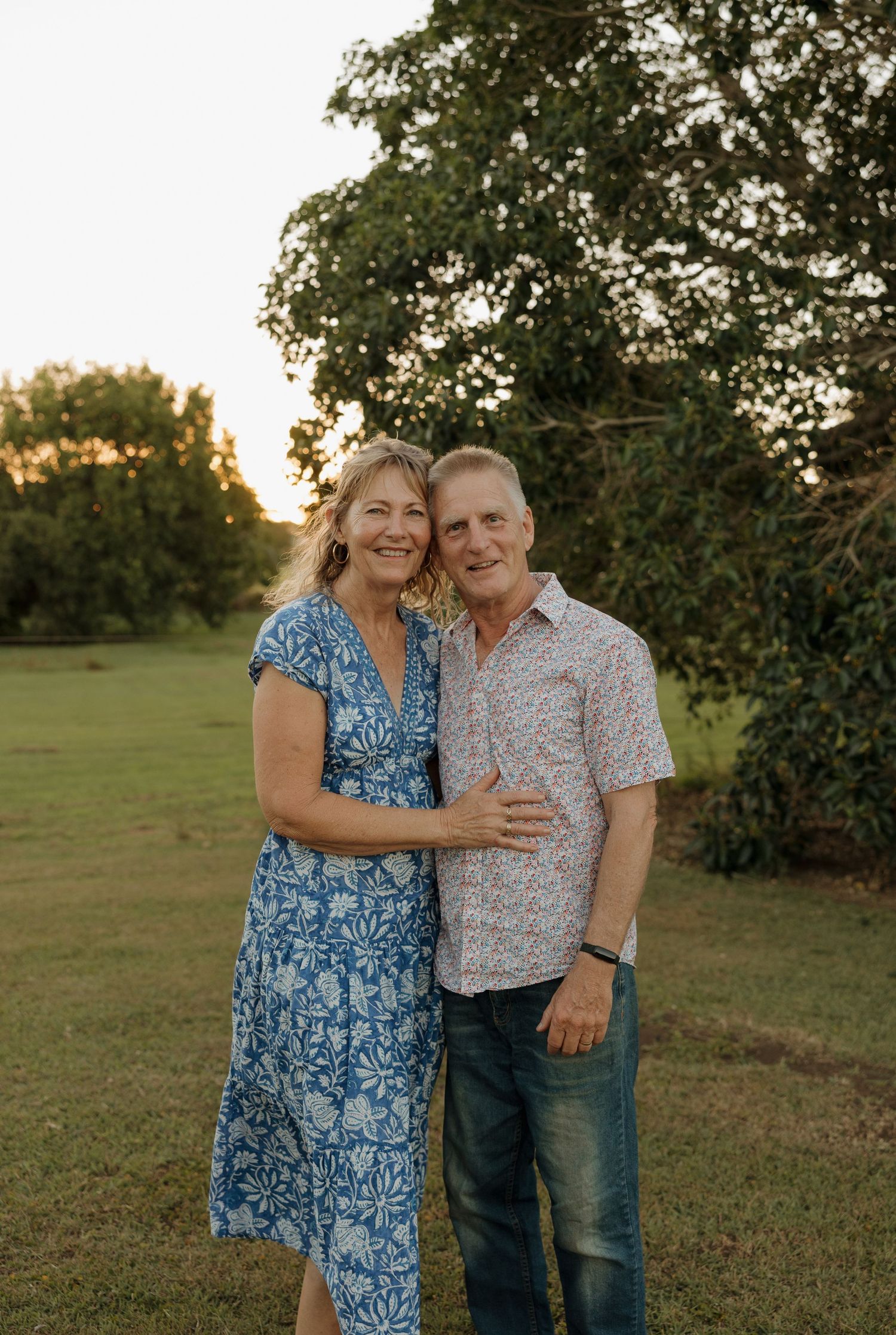 Smiling couple embracing outdoors at golden hour, woman in blue floral dress, lush green trees behind. Bundaberg Photographer