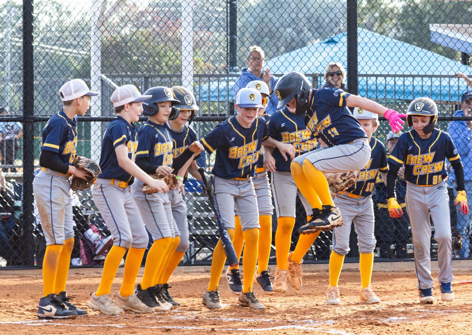 Baseball team sits along fence watching game from dugout in yellow uniforms.