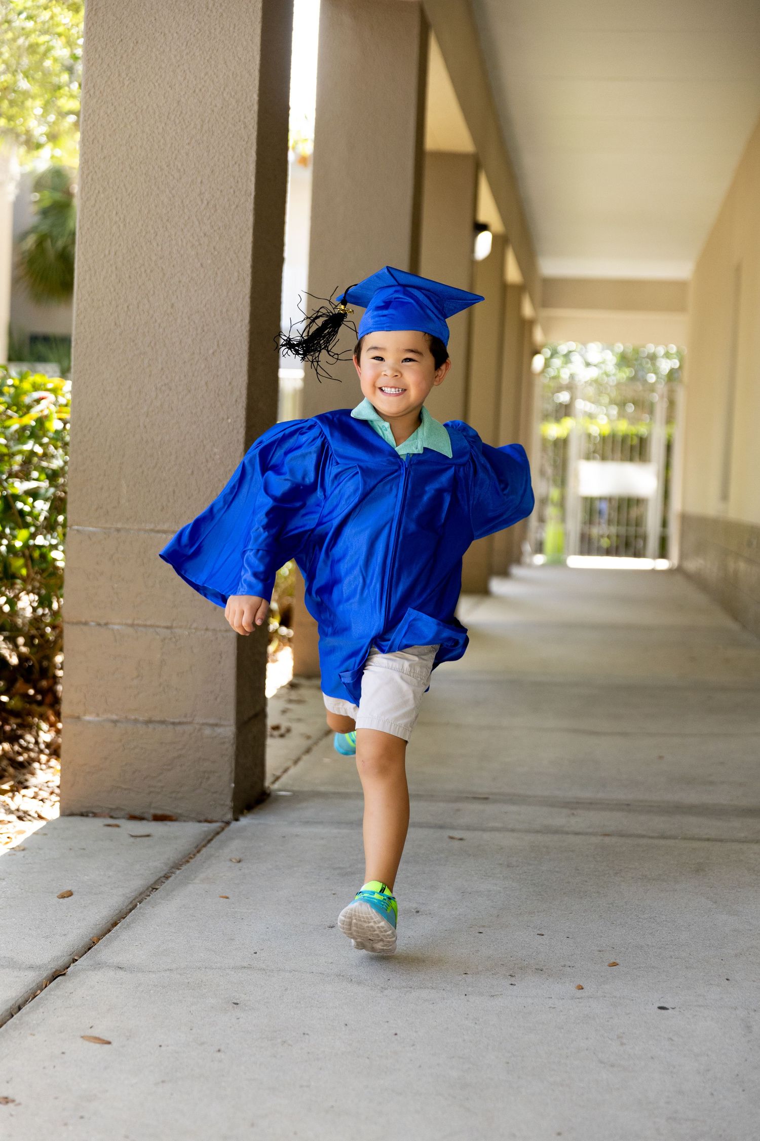 Young child joyfully running in blue graduation cap and gown along school corridor, celebrating academic milestone.