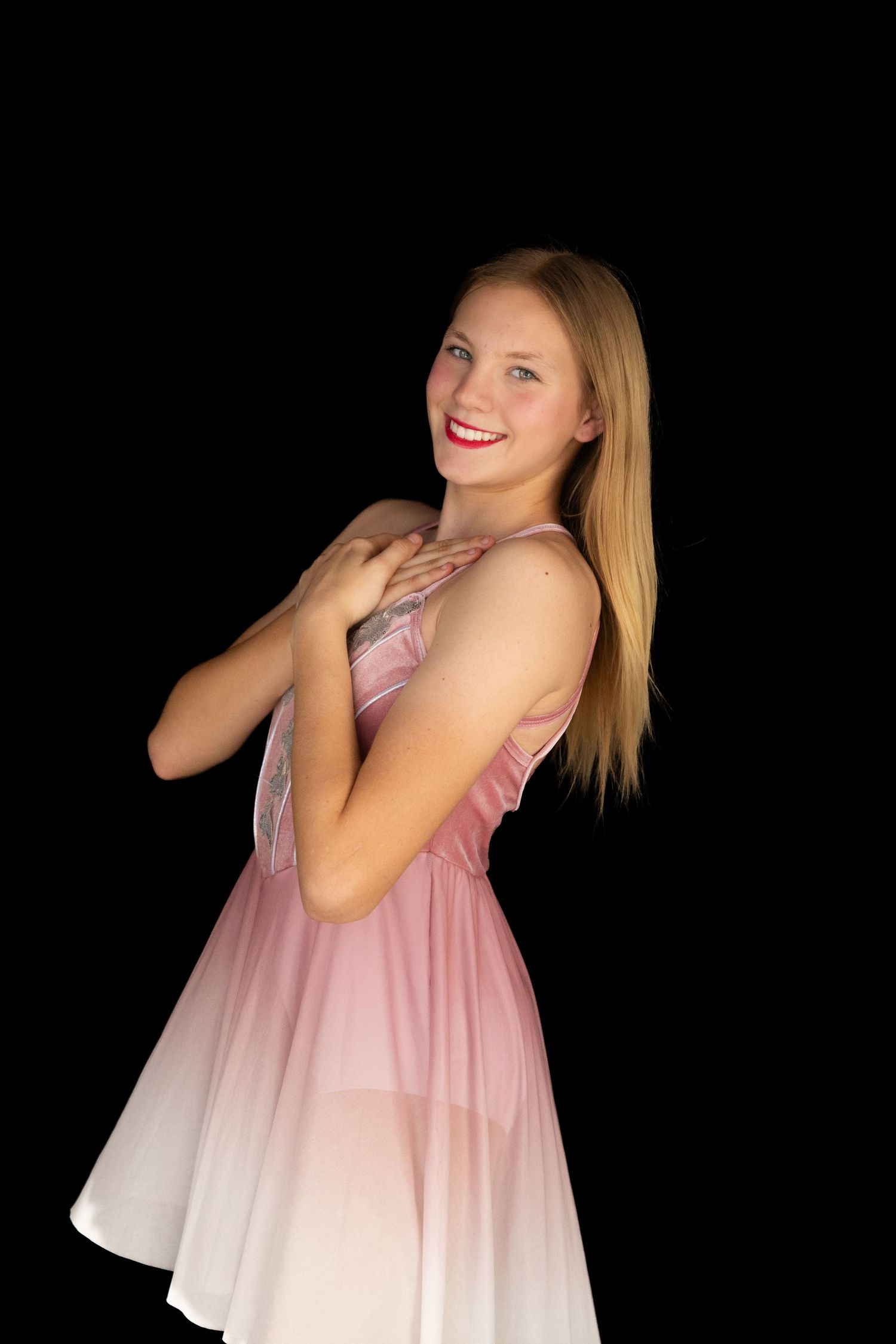 Smiling blonde teen girl in pink ombre dress posing against black background.