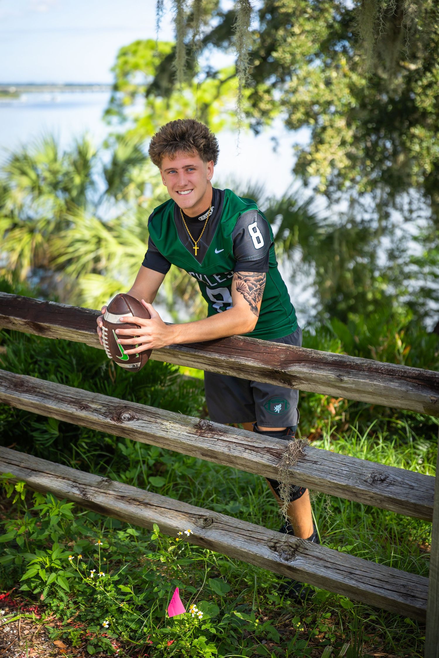 Young football player in green jersey #8 leaning on wooden fence holding football outdoors near waterfront.