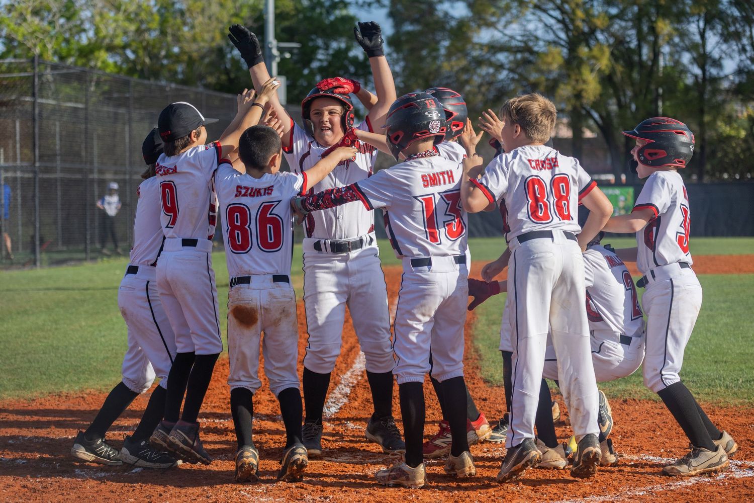 Youth baseball players celebrate together at home plate during a game, wearing white and red uniforms.