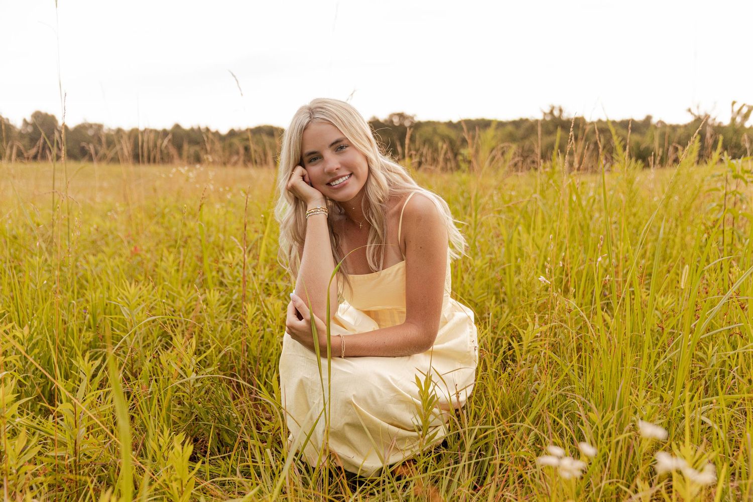 A smiling blonde woman in a yellow dress sits in a lush green meadow field on a bright day.