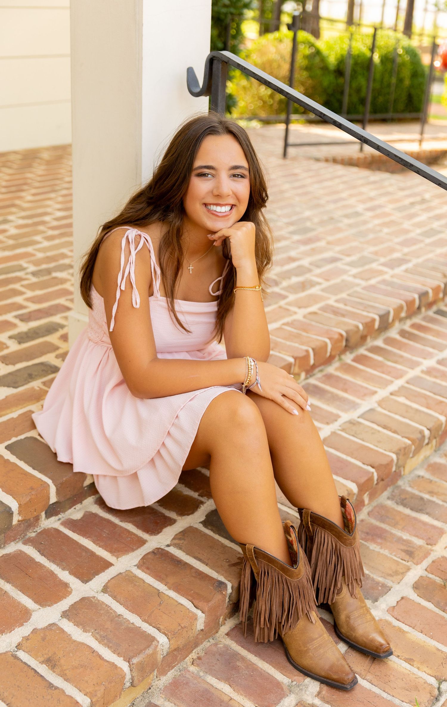Young woman in pink dress and brown fringe cowboy boots sitting on brick steps, smiling cheerfully.