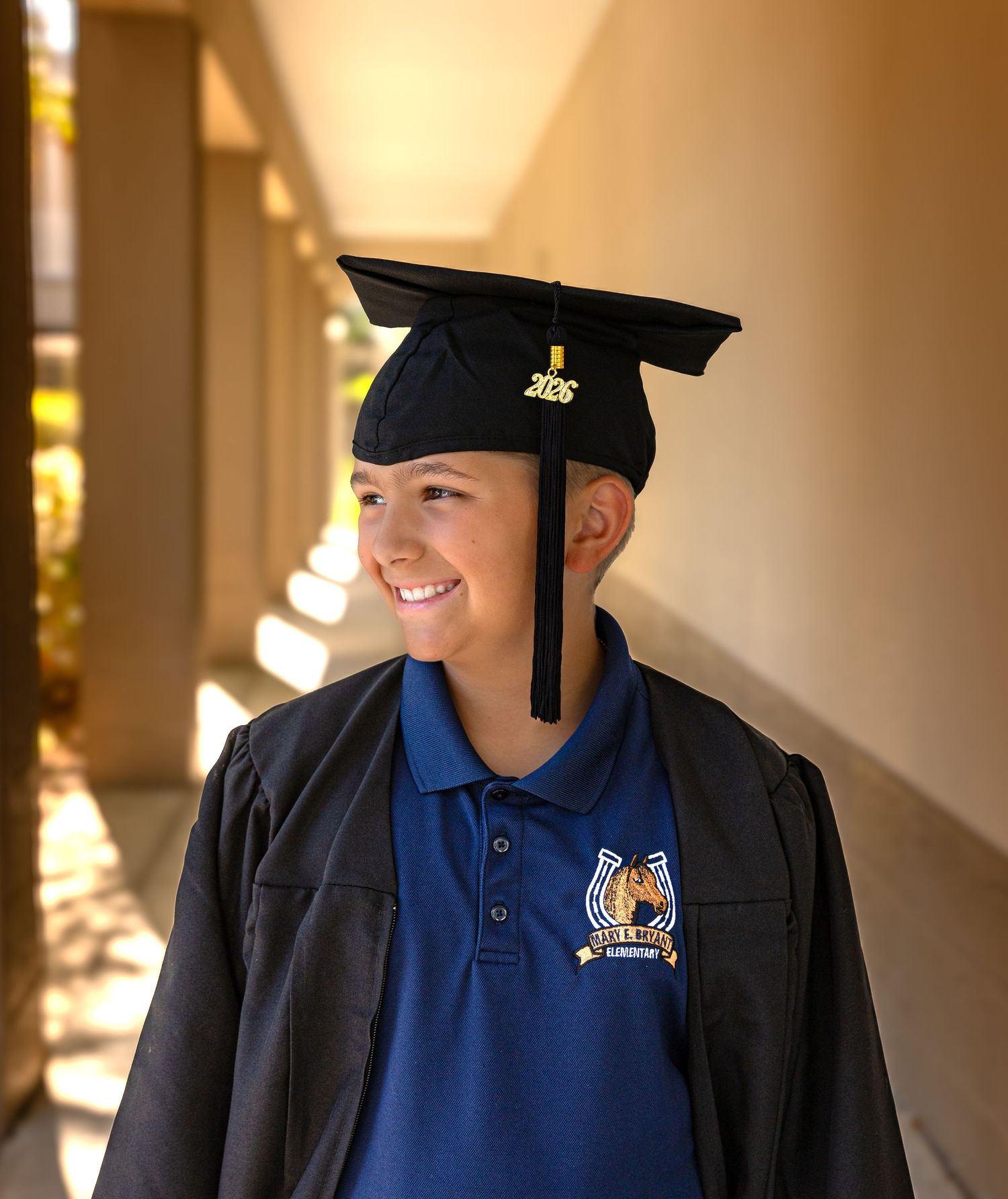 Smiling student wearing black graduation cap and gown over blue polo shirt, standing in sunlit corridor.