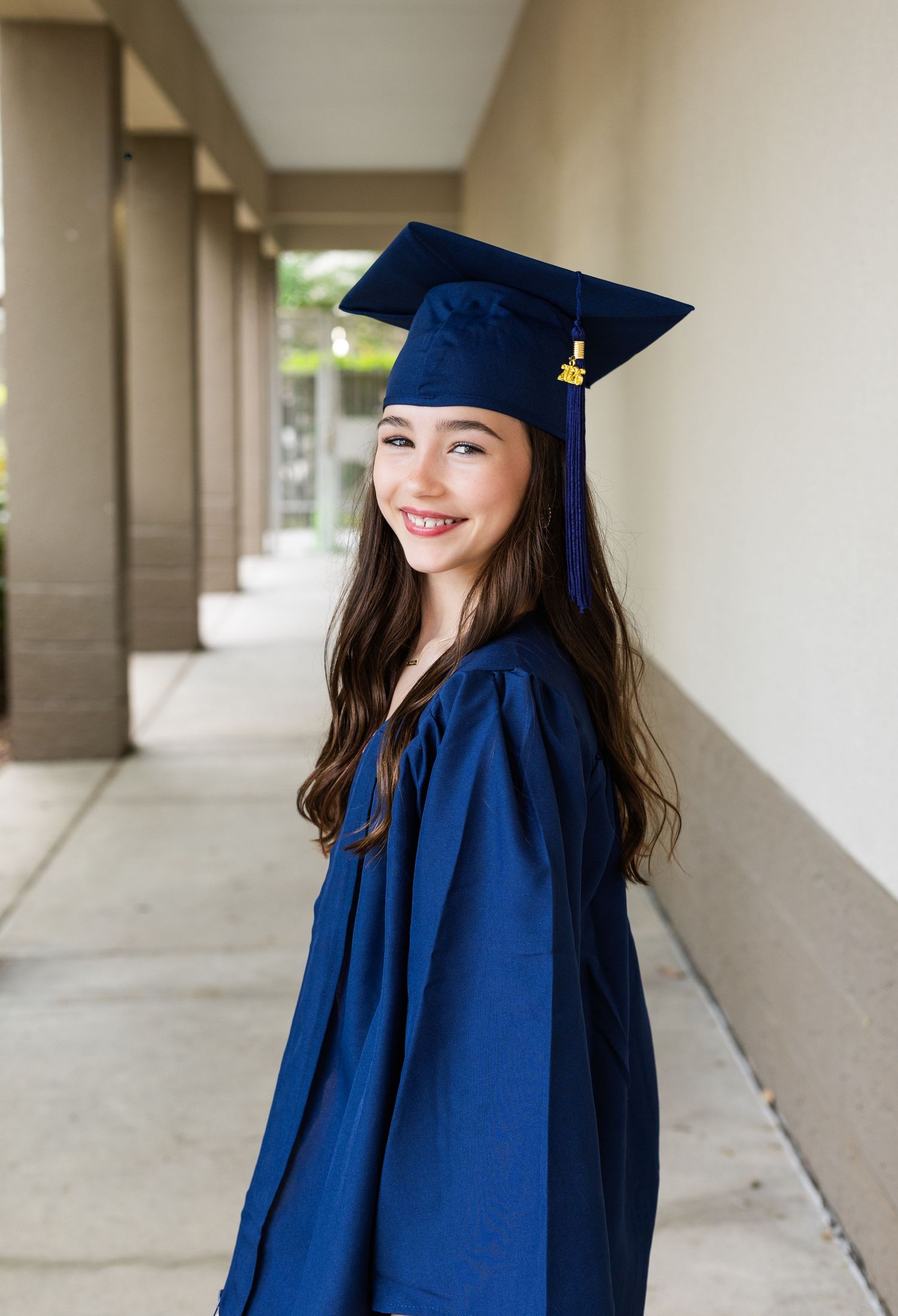 Smiling young girl in navy blue graduation cap and gown posing in a hallway colonnade.