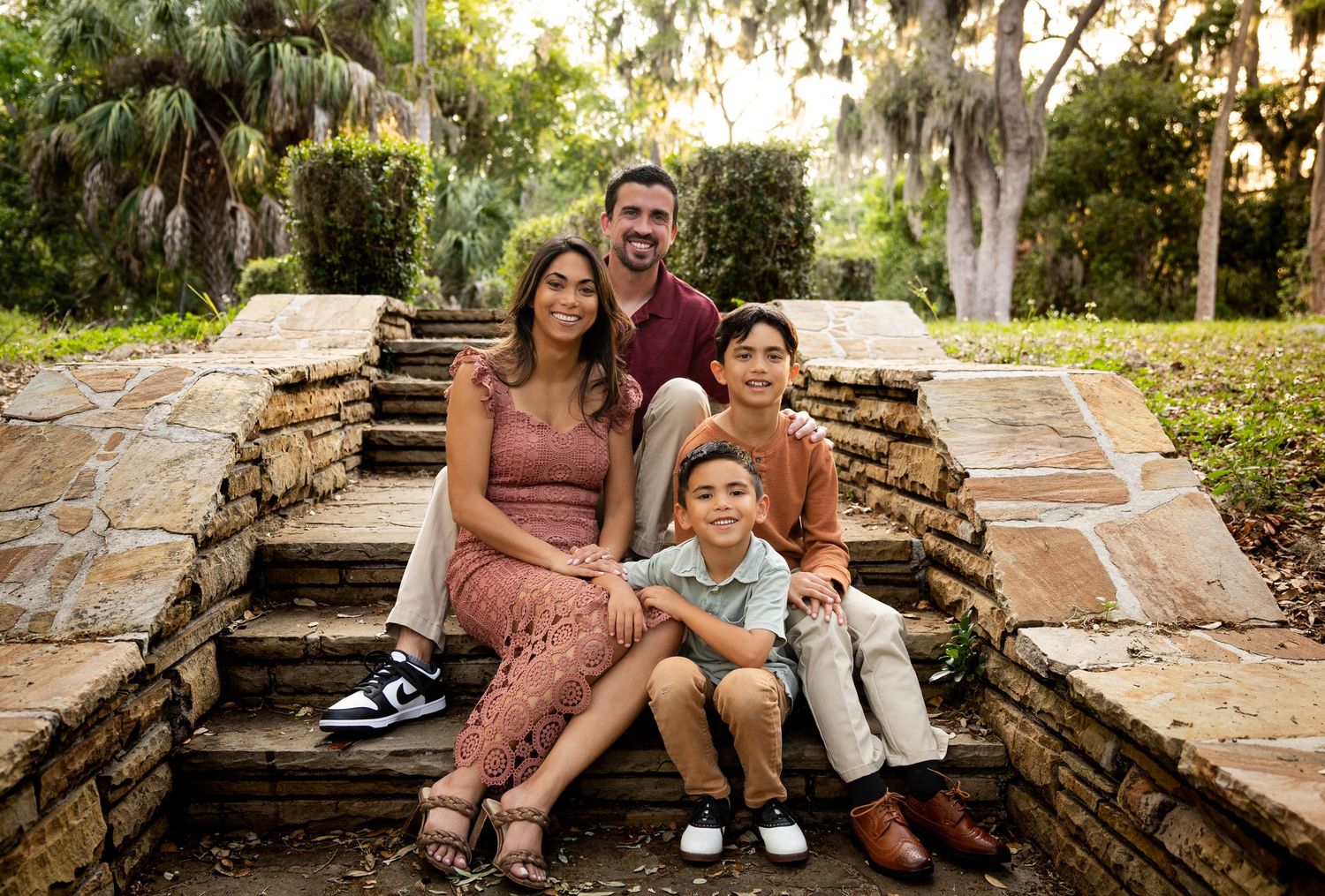 Family of four smiling on stone steps surrounded by lush greenery in an outdoor park setting.
