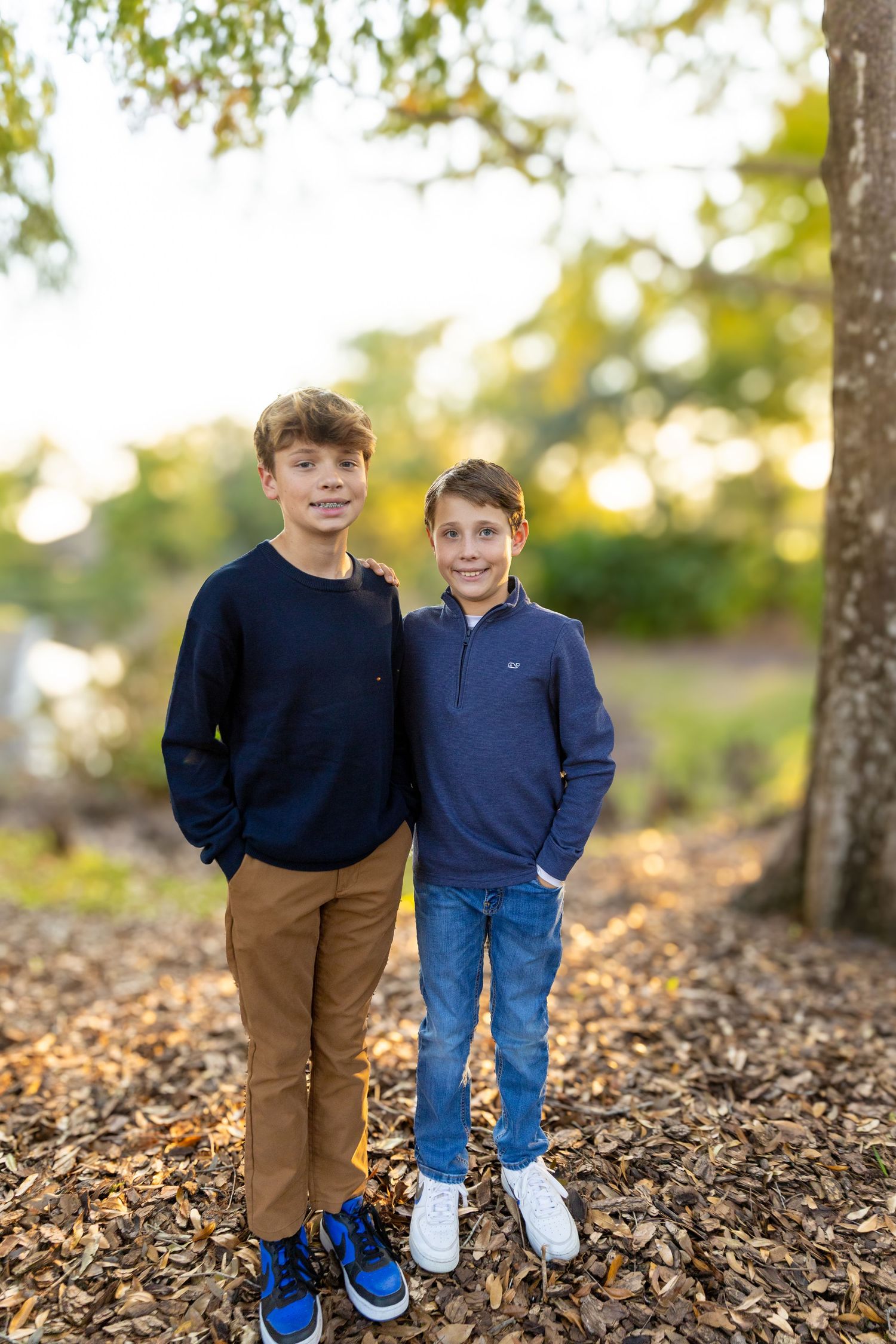 Two young boys in navy sweaters stand together on fallen leaves during autumn photoshoot.
