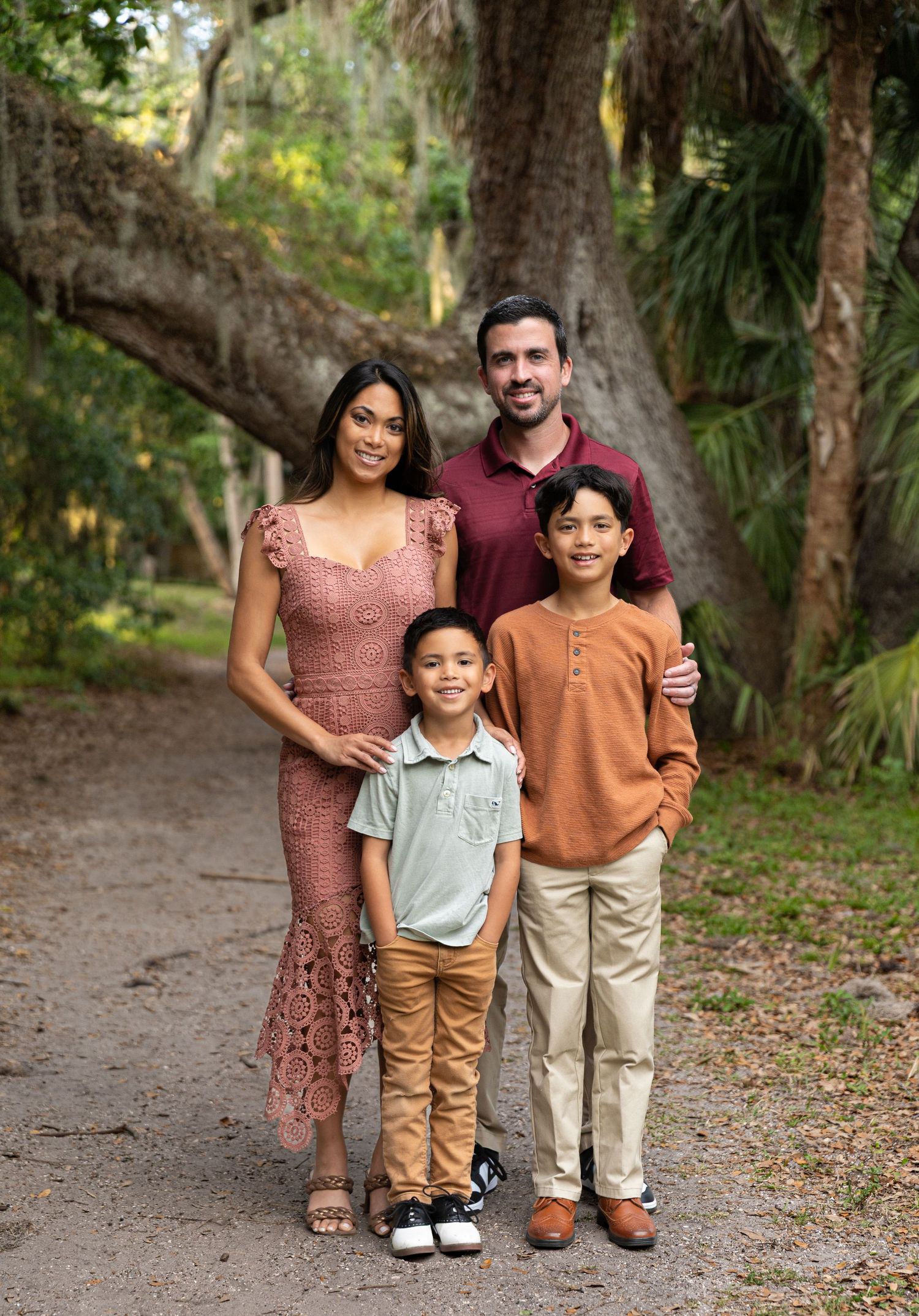 Family of four posing on a scenic tree-lined path, parents and two boys smiling in coordinated outfits.