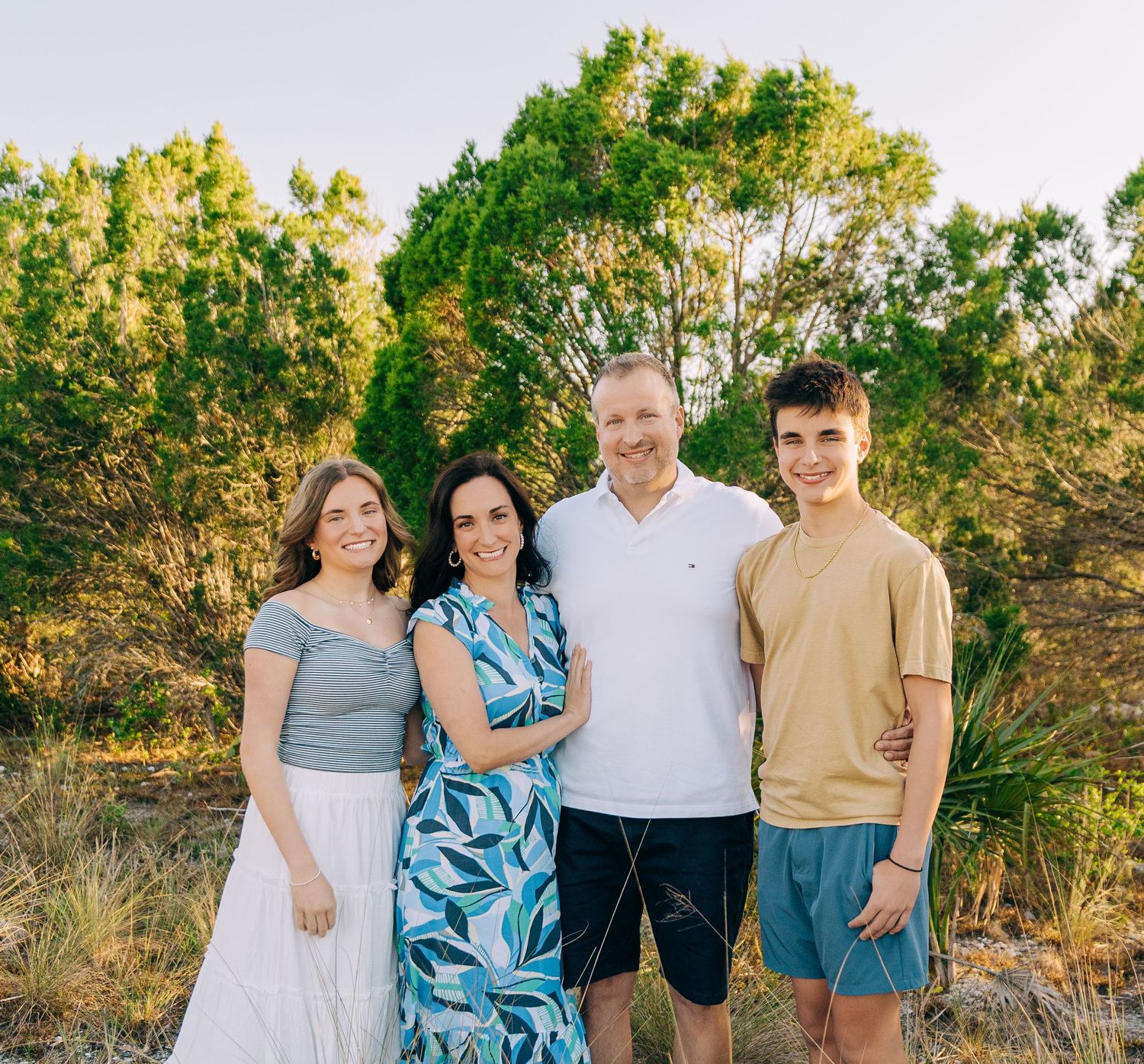 Happy family of four smiling outdoors surrounded by lush green trees in a sunny natural setting.
