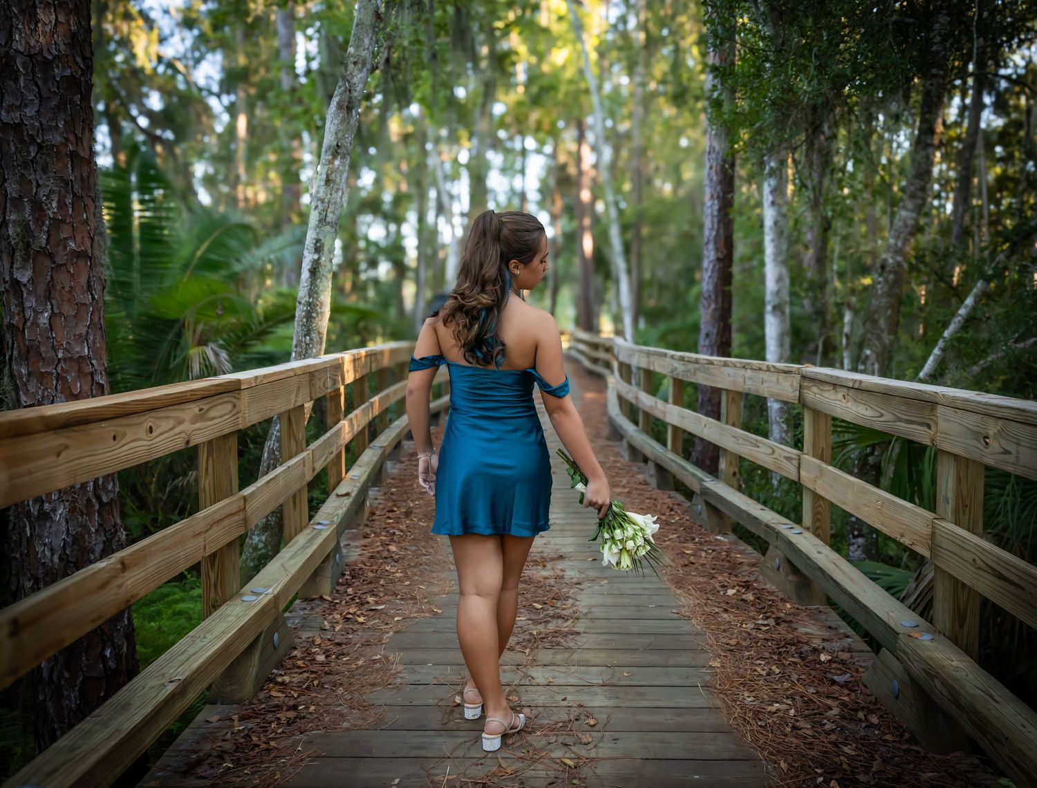 Woman in teal dress holding flowers walks along wooden boardwalk path through lush green forest.
